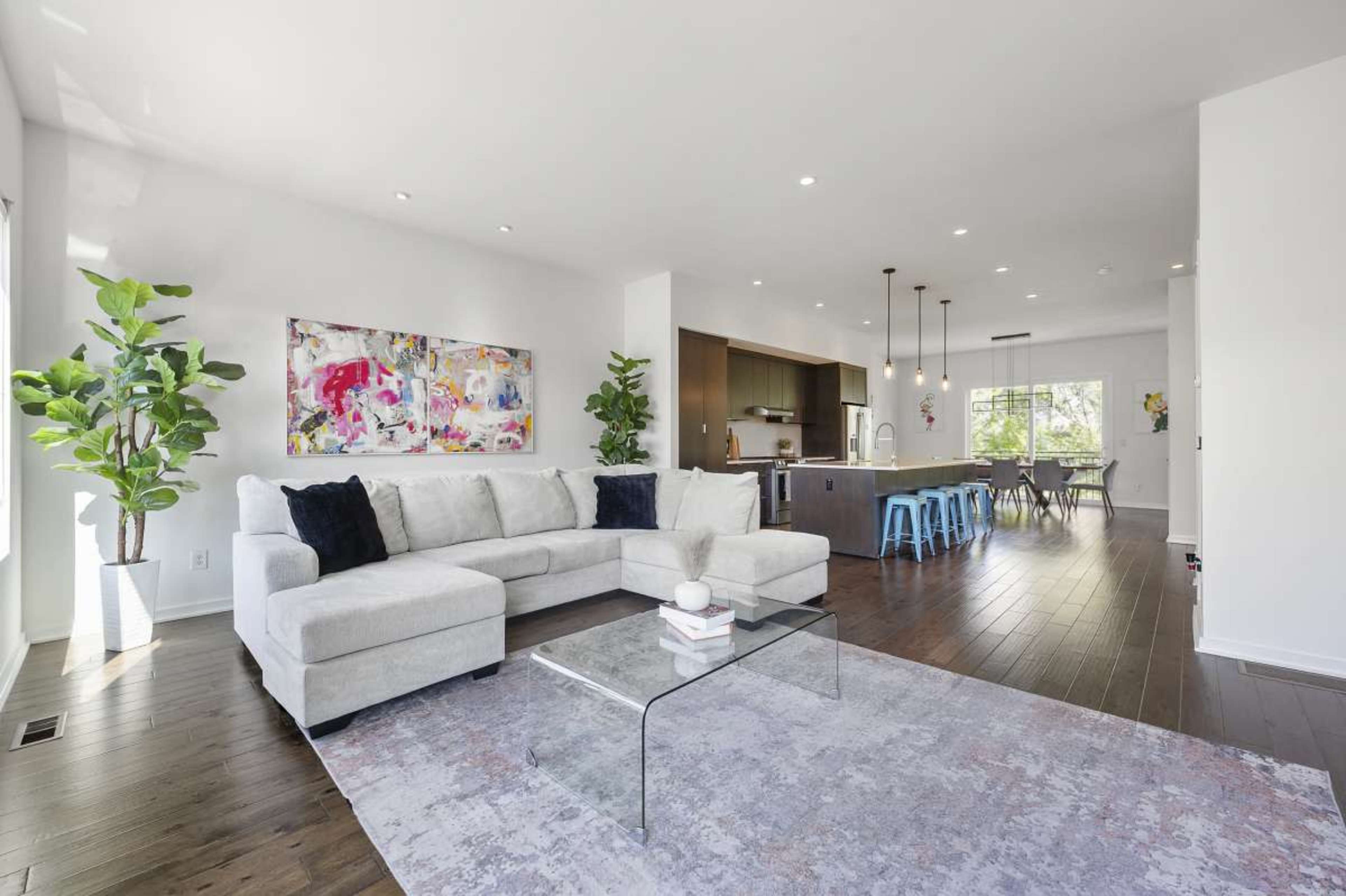 The image shows a modern living space with a light-colored sectional sofa, a glass coffee table, and an open kitchen area featuring barstool seating and dining space.