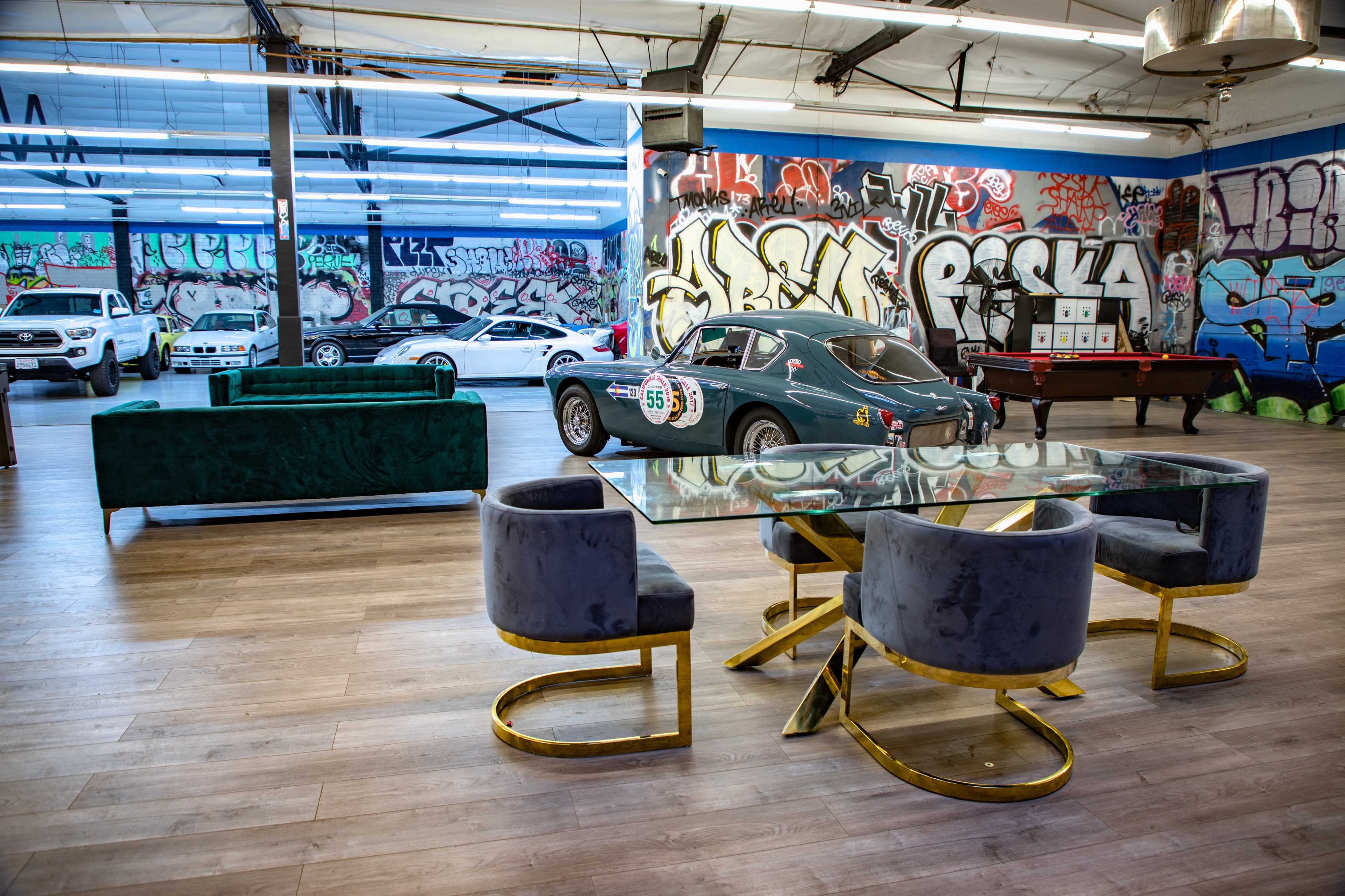 A spacious room featuring a glass-top dining table with curved chairs, vintage cars, and graffiti-covered walls.