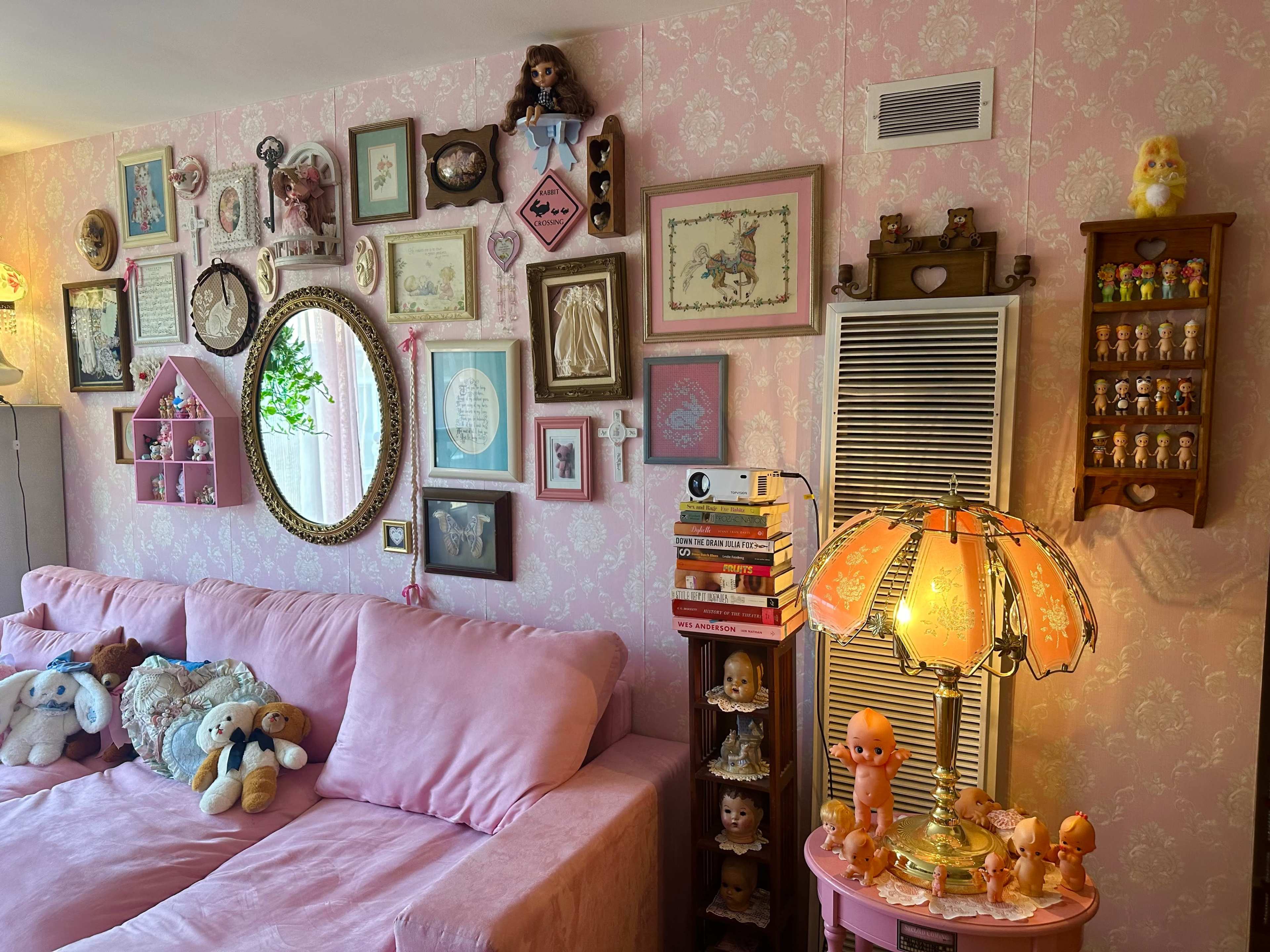 The scene features a pink-themed living room wall adorned with various framed artworks, vintage dolls, and a decorative lamp beside a plush pink couch.