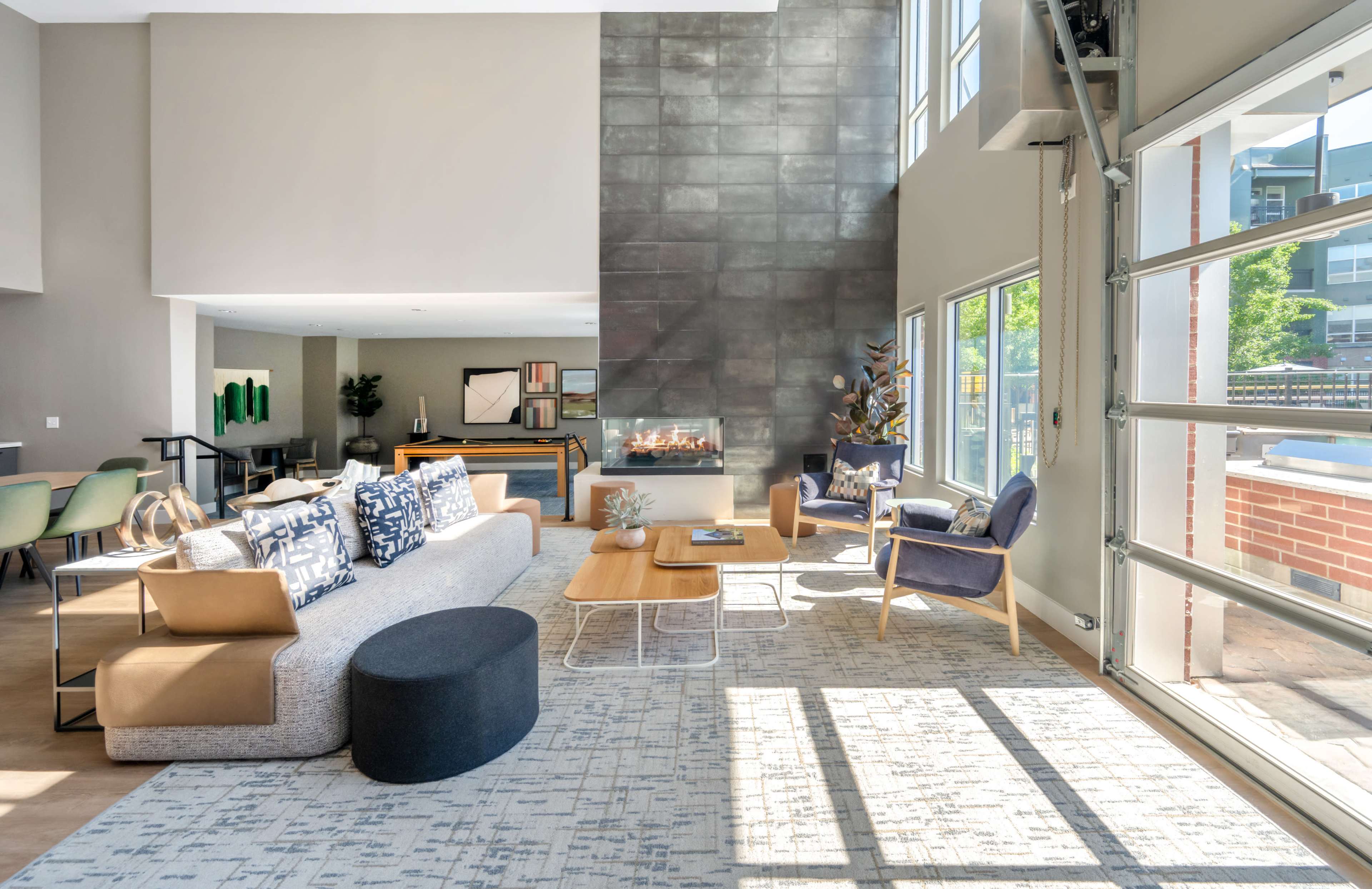 The image shows a modern living space featuring a stylish sectional sofa, a coffee table, and large windows allowing natural light to fill the area, with a fireplace positioned against a textured wall.