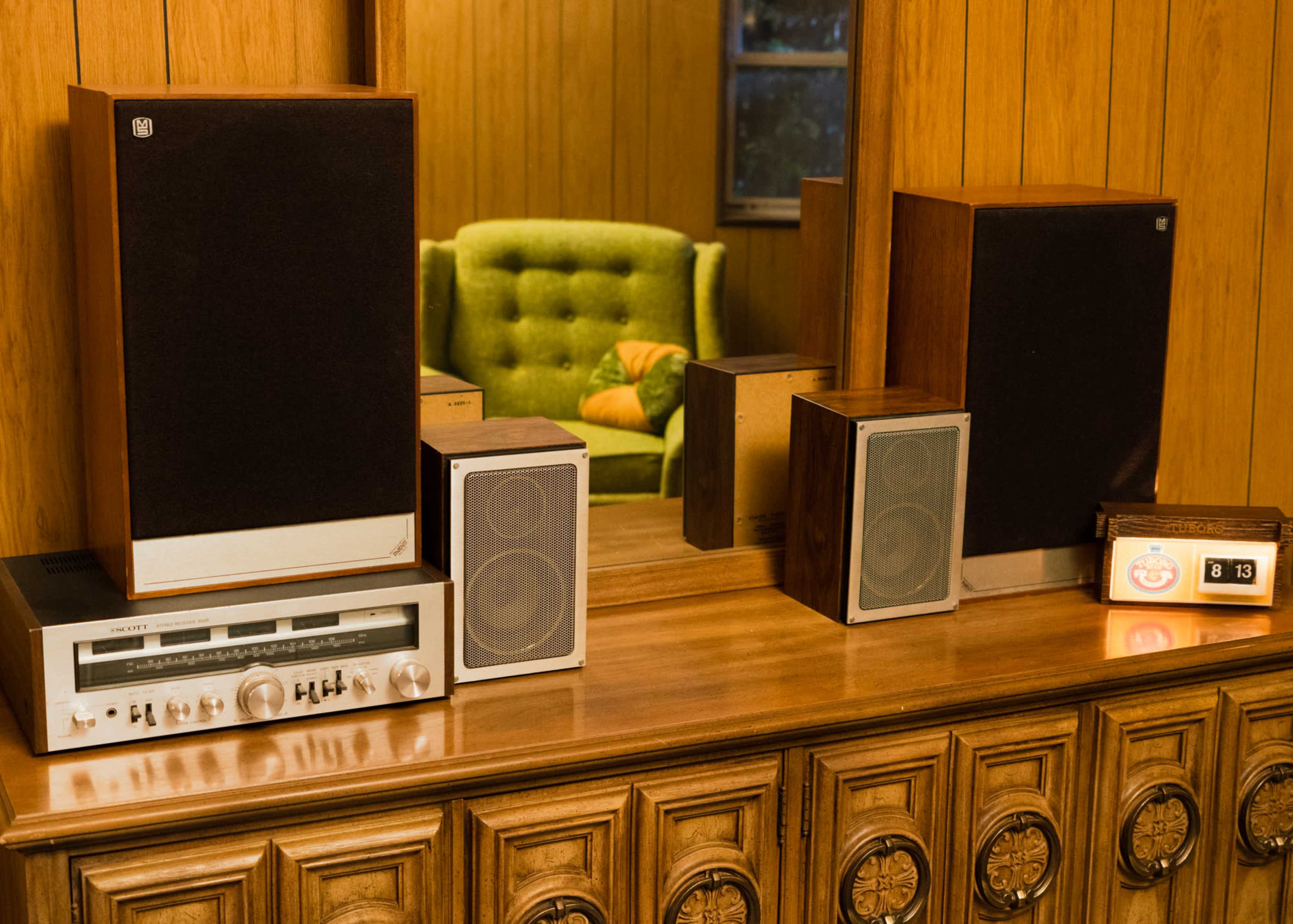 The image shows a wooden sideboard displaying vintage audio equipment, including speakers and a receiver, alongside a green upholstered chair in a room with wood paneling.