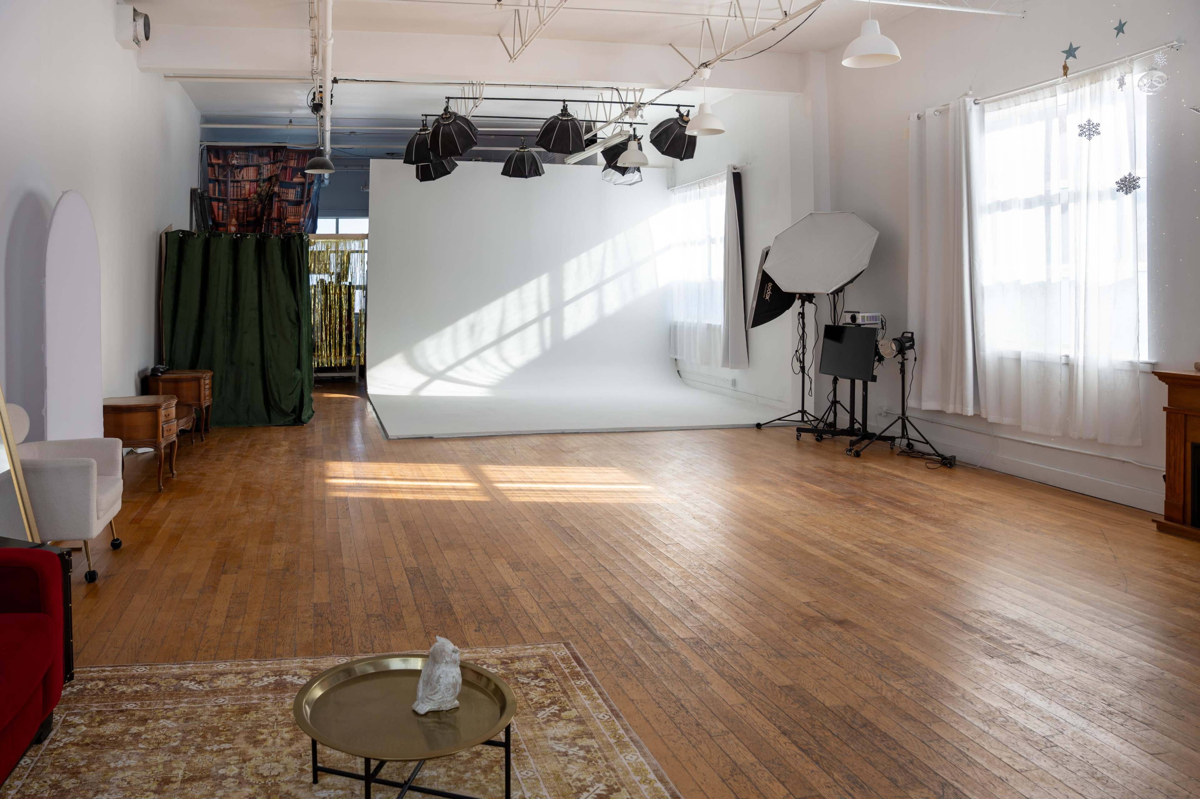 The image shows a spacious, well-lit photography studio with wooden floors, a white backdrop, and various lighting equipment set up around the area.