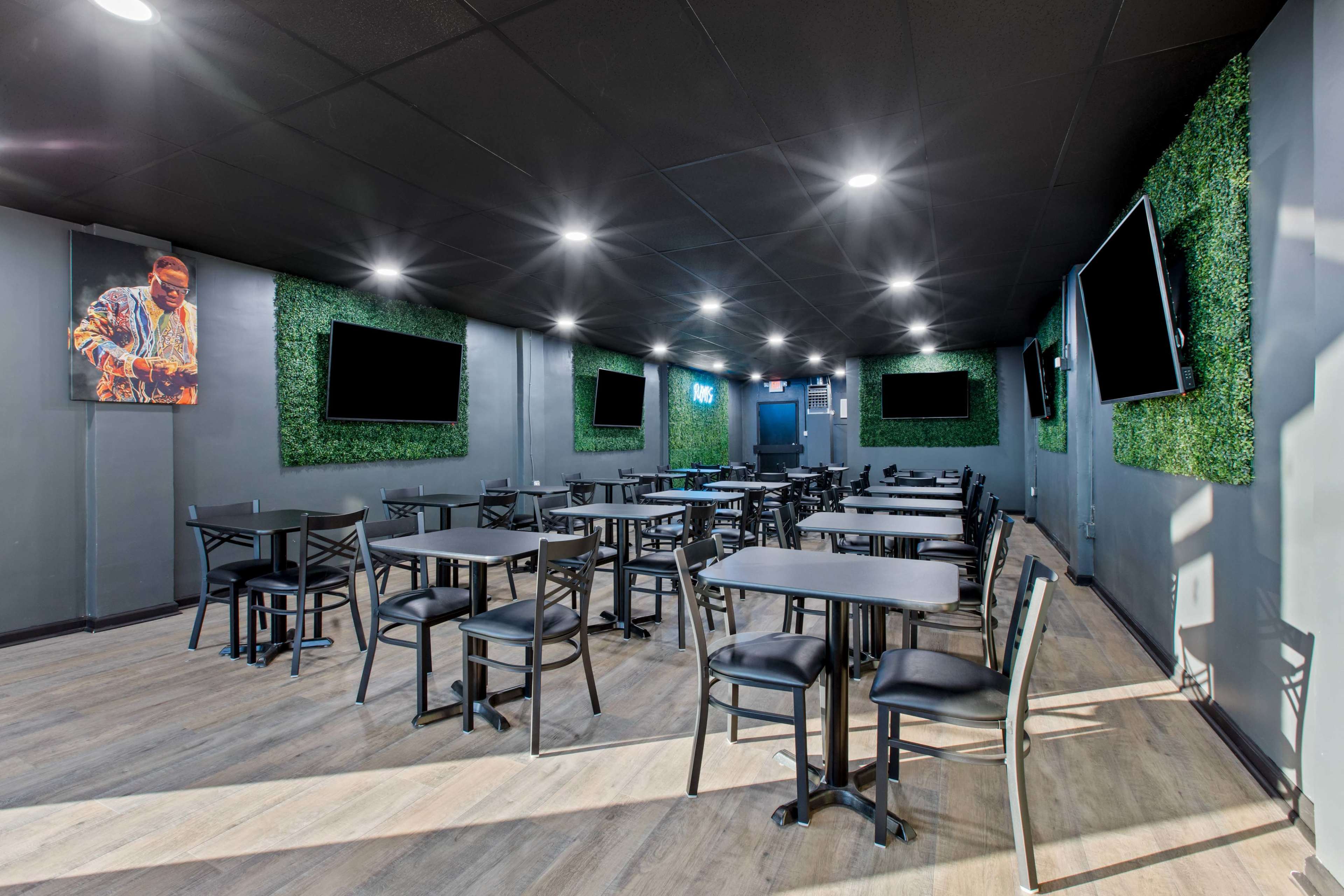 An empty dining area with black tables and chairs, artificial green wall panels, and multiple flat-screen TVs mounted on the walls.