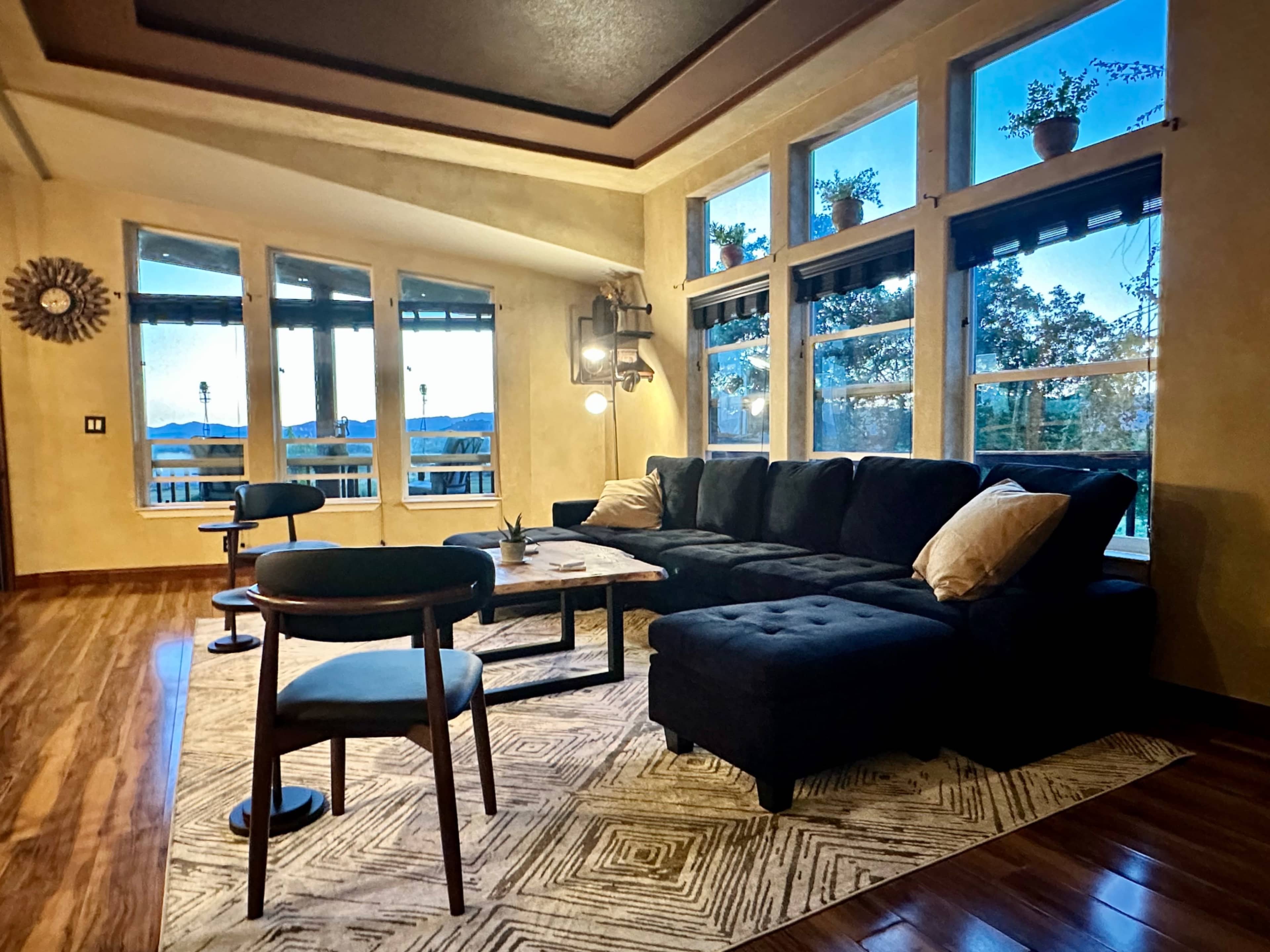 The image shows a spacious living room with large windows, a sectional couch, a coffee table, and a patterned rug.