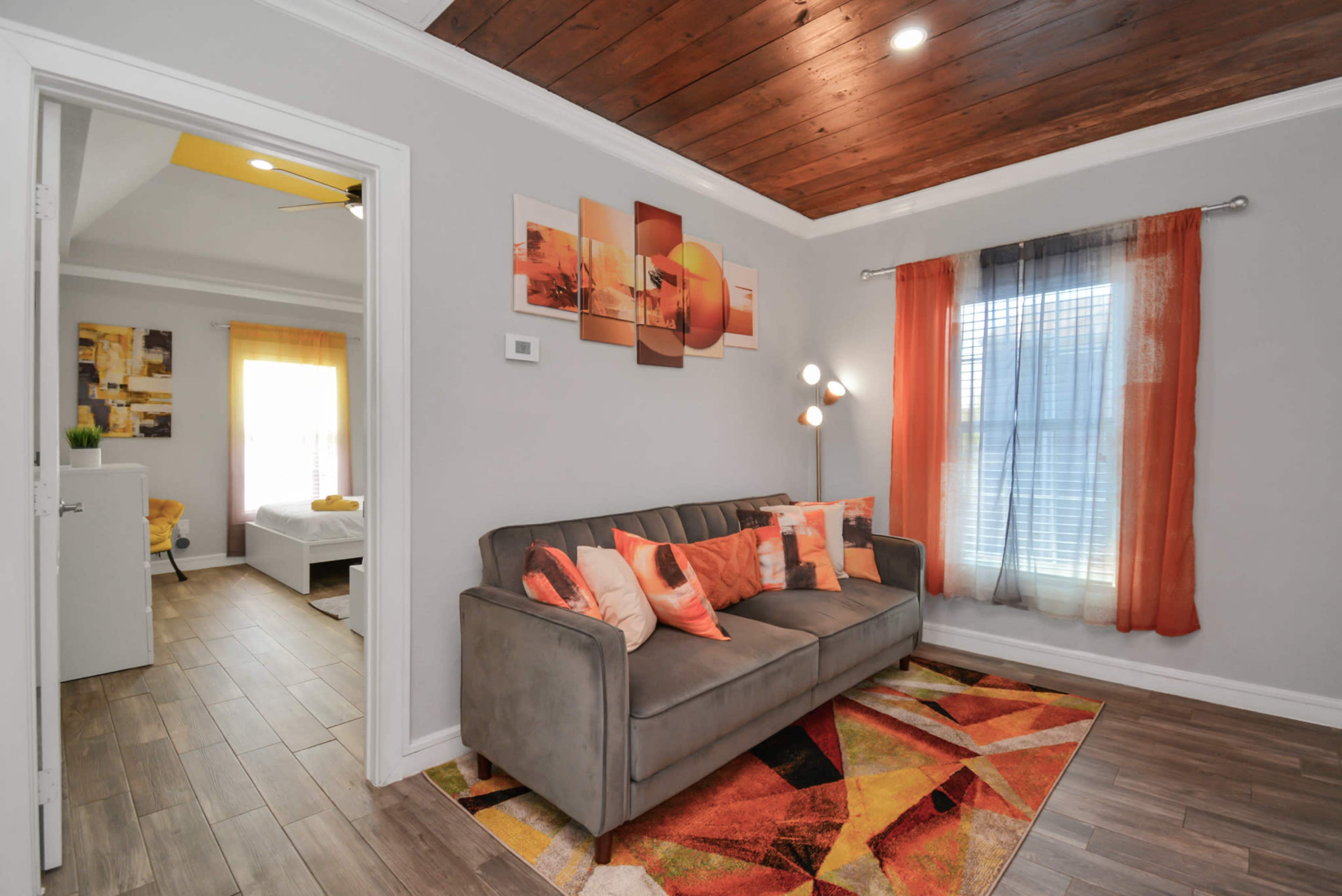 A living room with a gray sofa, colorful throw pillows, a geometric rug, and a window with sheer curtains, leading into another room.
