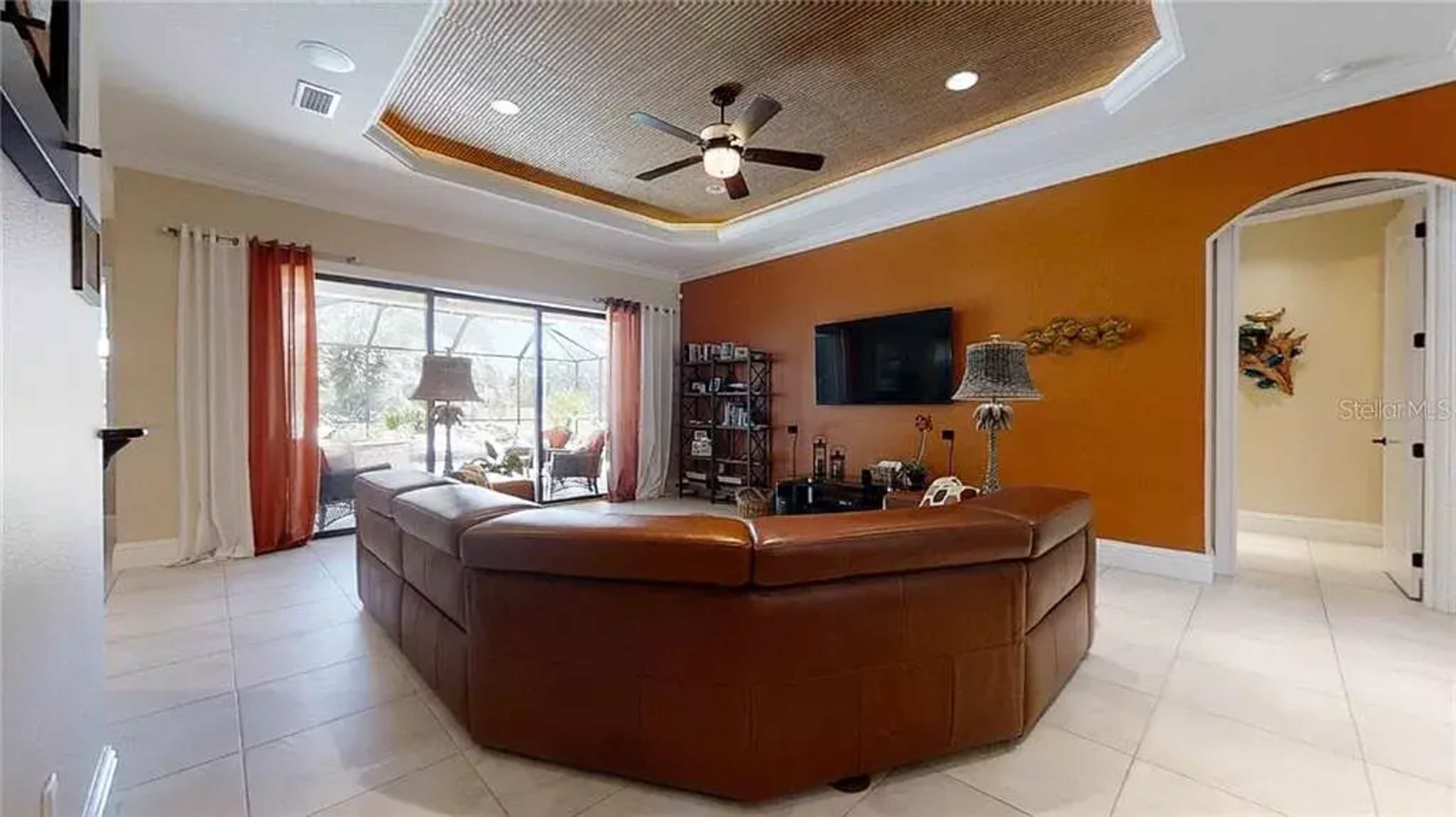A living room with a brown sectional couch, a television mounted on the wall, and large windows overlooking a patio area.