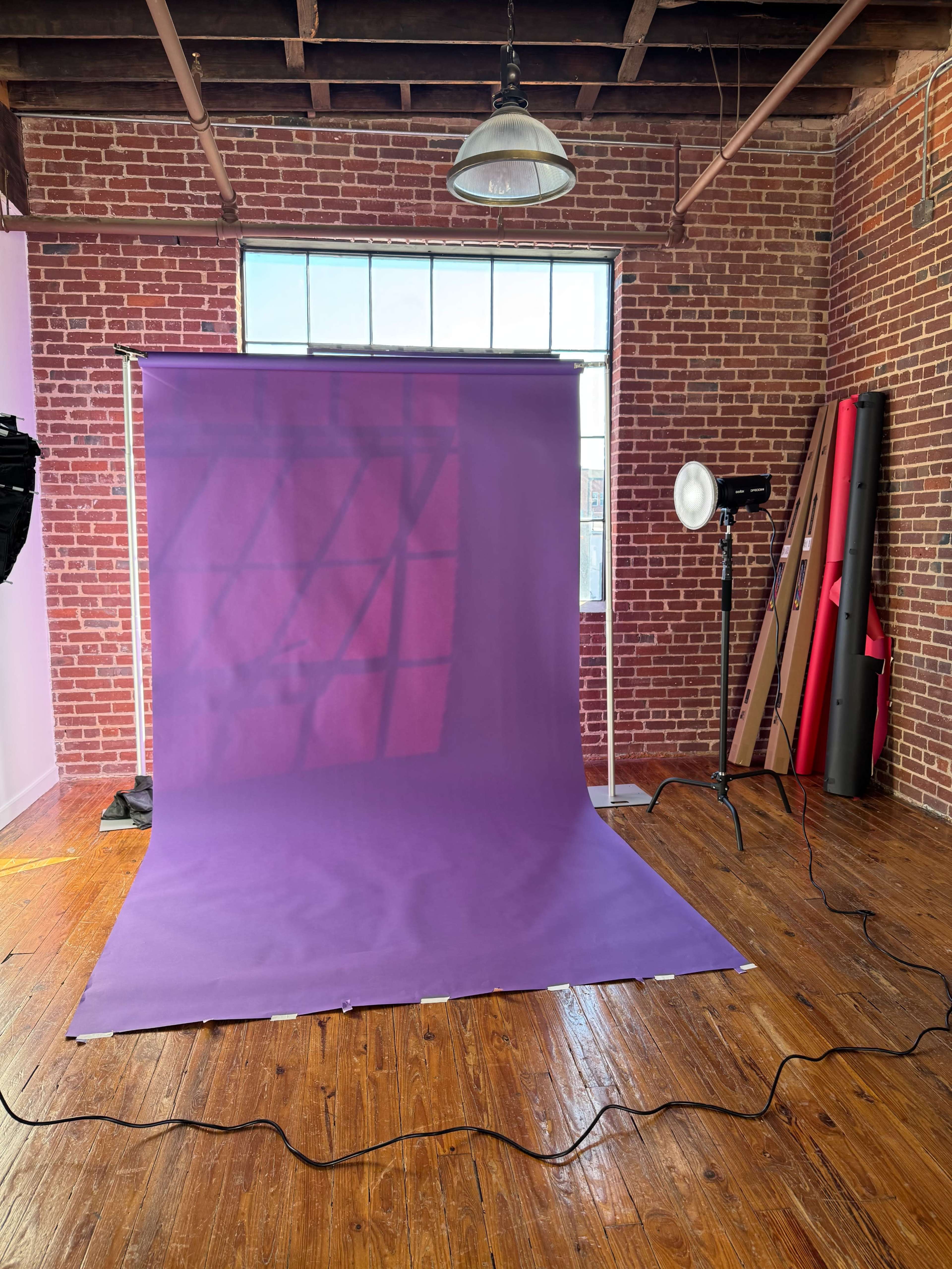 A spacious studio with a purple backdrop, wooden floors, and various lighting equipment is set up against a brick wall.