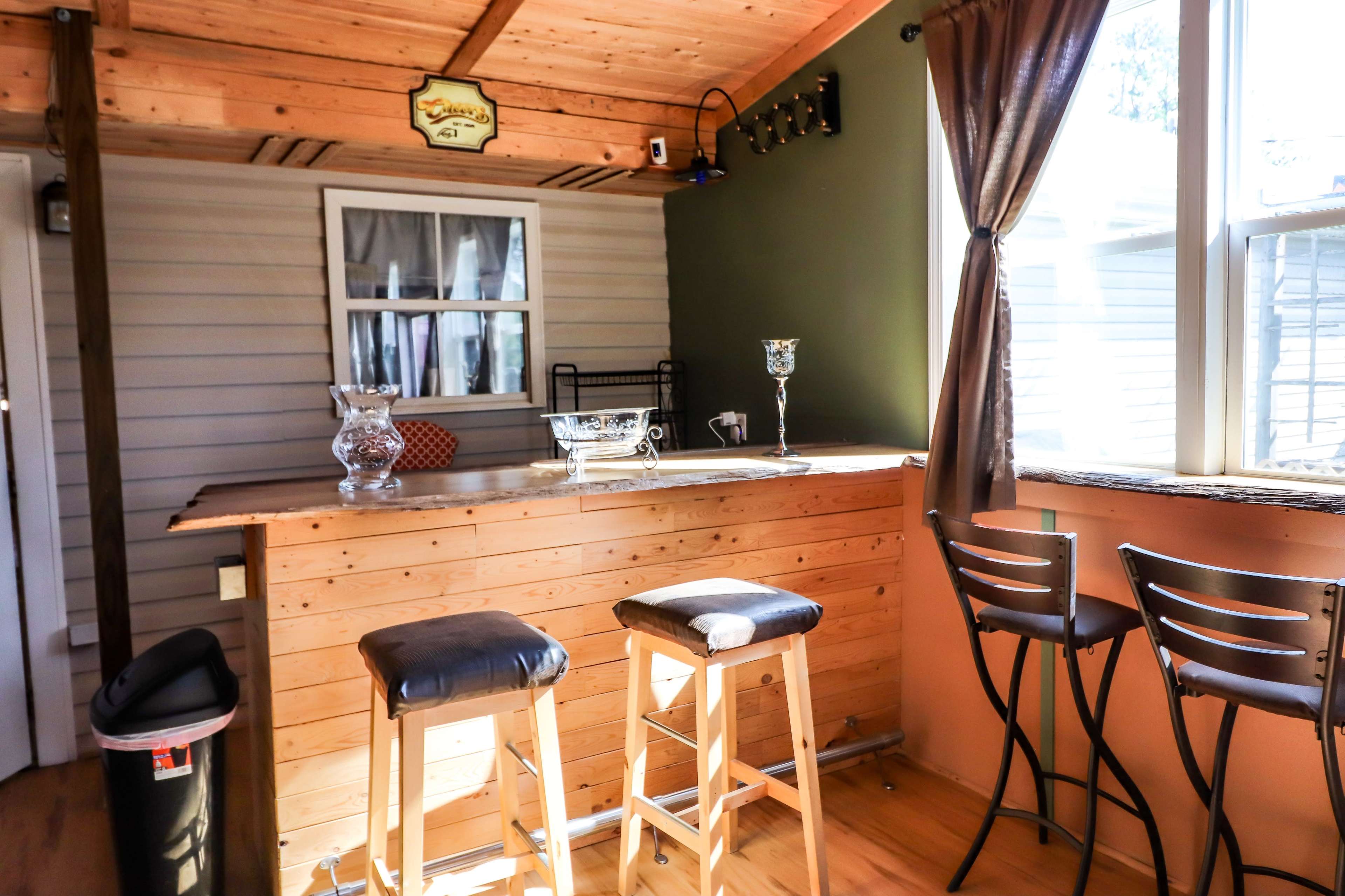 The image shows a cozy home bar area featuring a wooden bar counter with three stools and a window that allows natural light to enter the space.