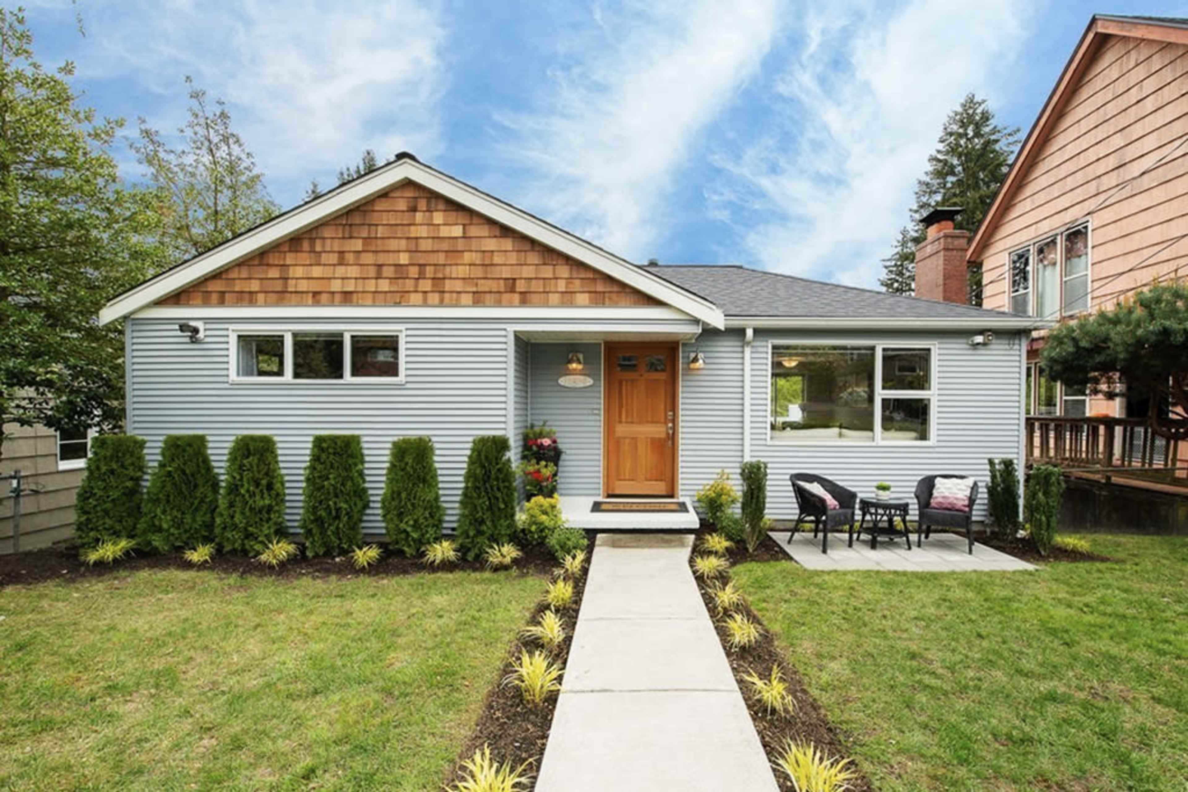 A single-story house with a light blue exterior, a wooden front door, and a neatly landscaped yard featuring a pathway and manicured plants.