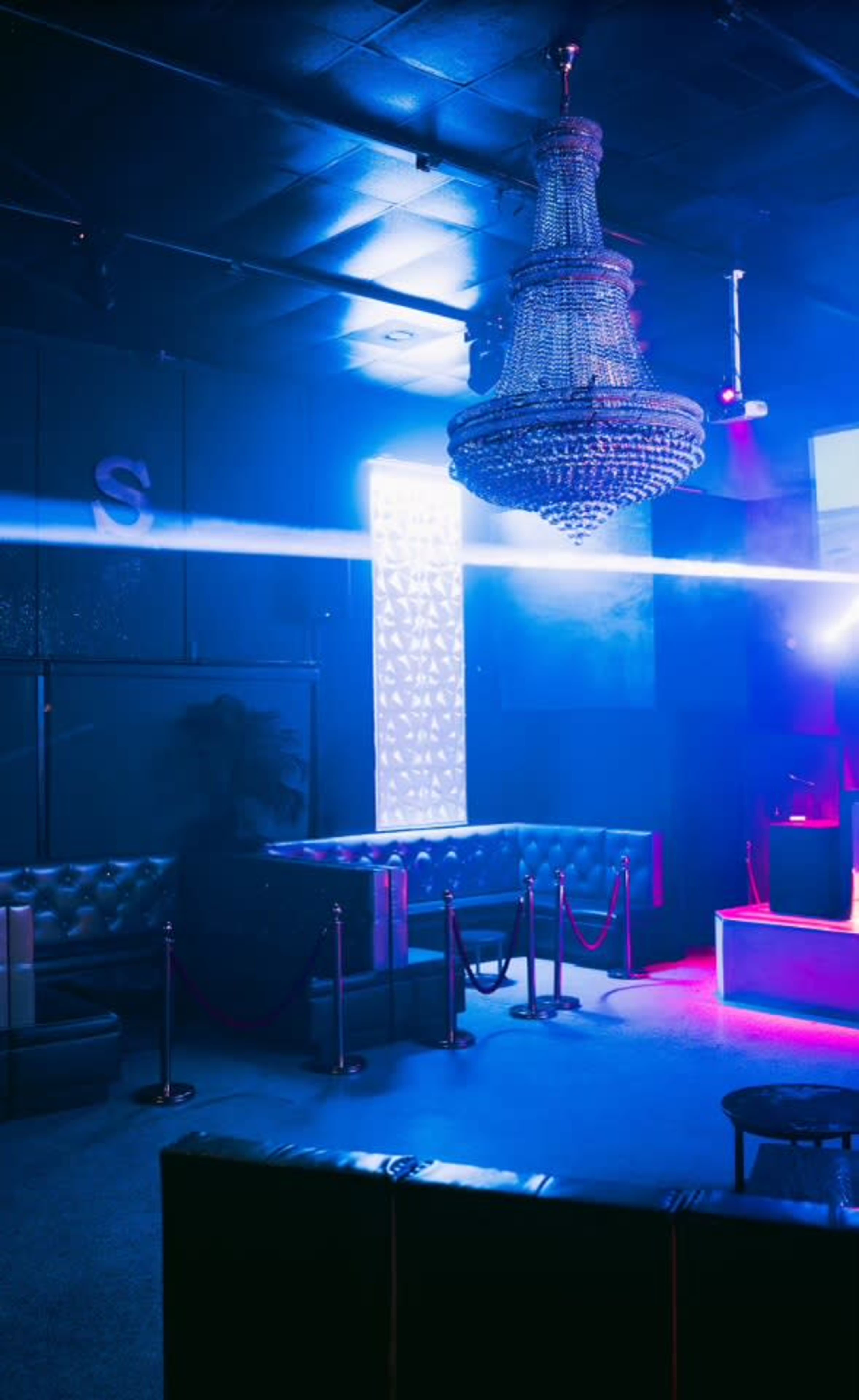 A dimly lit nightclub features a crystal chandelier, velvet seating, and illuminated walls with a colorful light display.