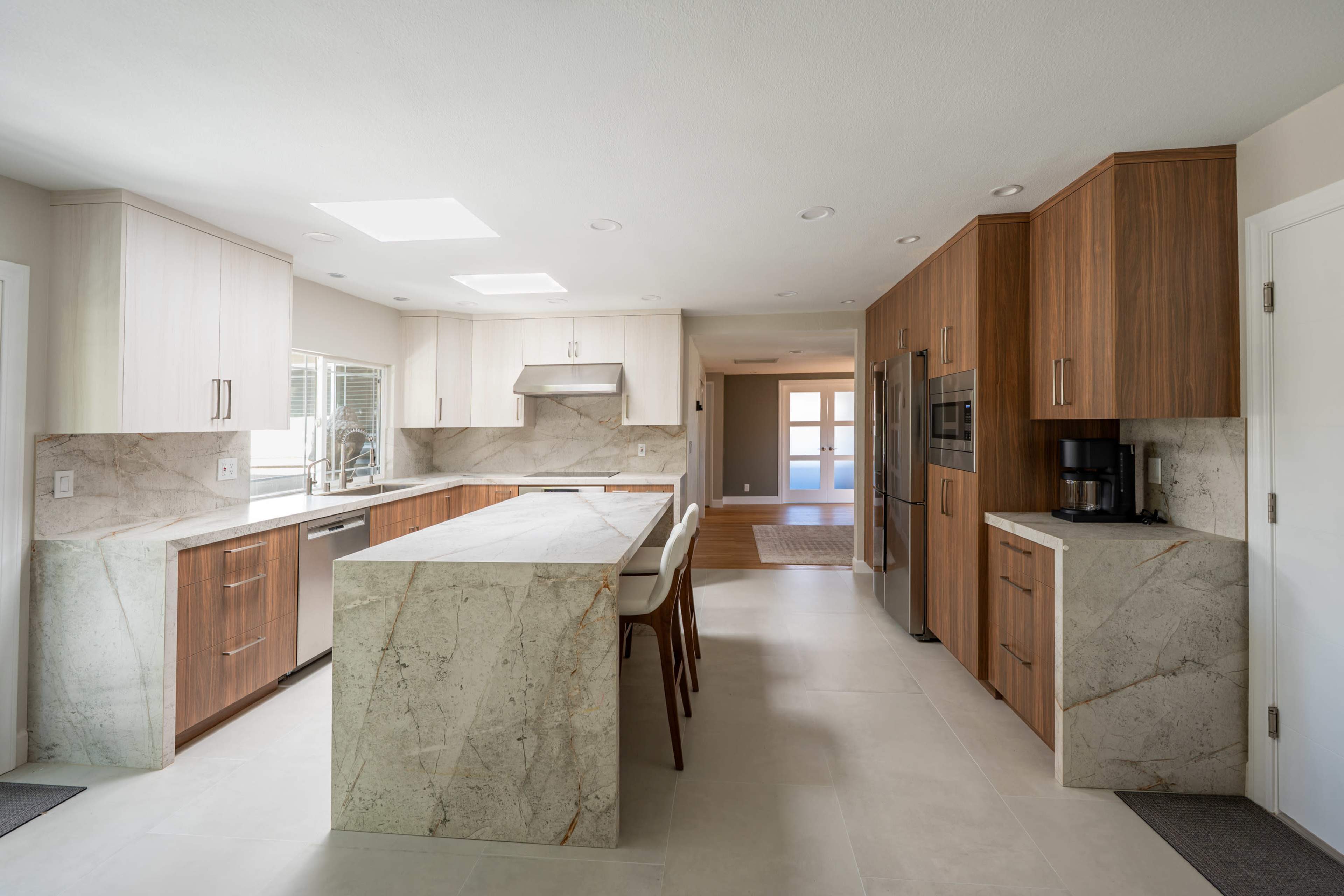 The image shows a modern kitchen featuring a large island with marble countertops, wooden cabinetry, and stainless steel appliances.