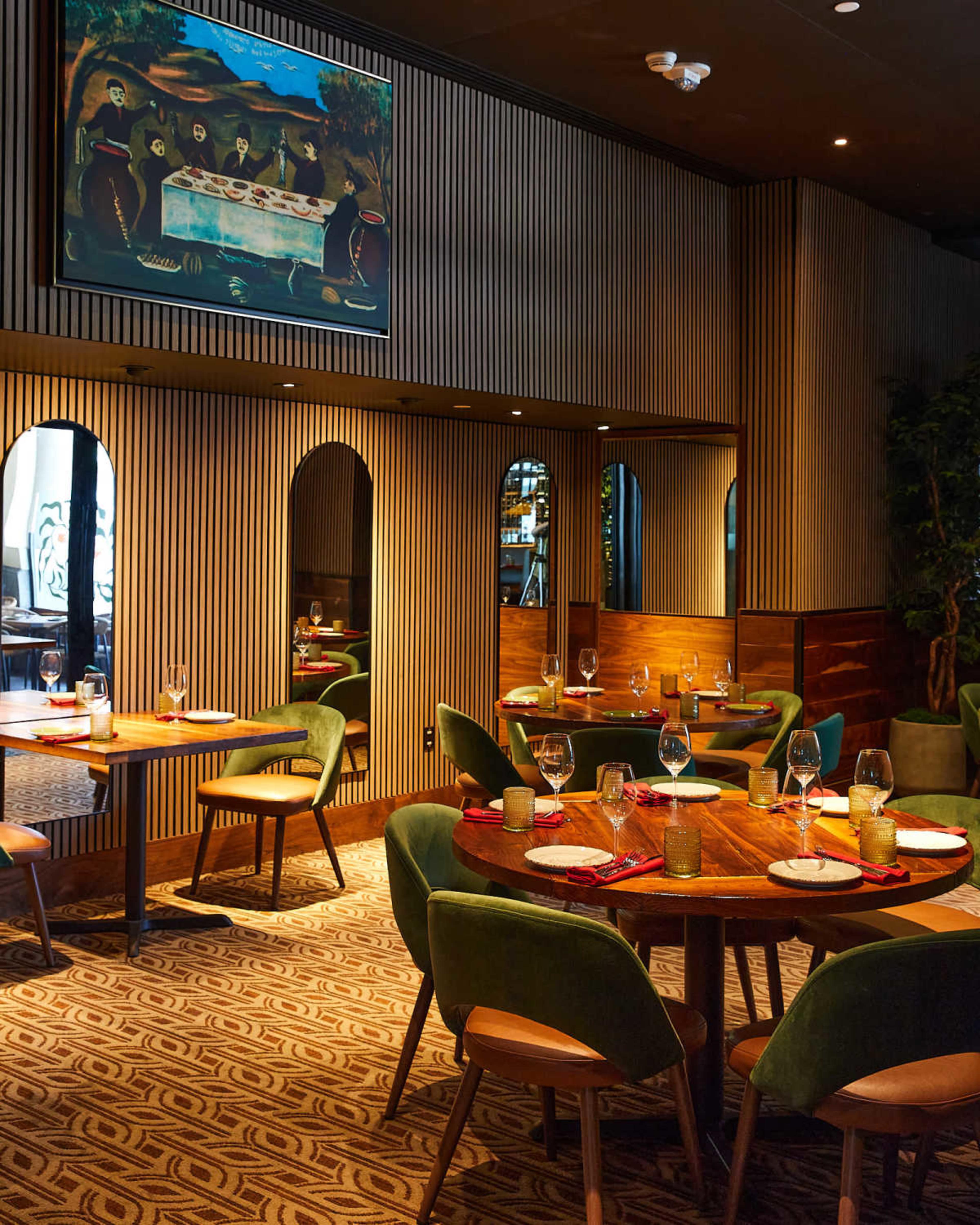 The image shows a modern restaurant interior with wooden furniture, green upholstered chairs, and a painting on the wall, set with tables for dining.