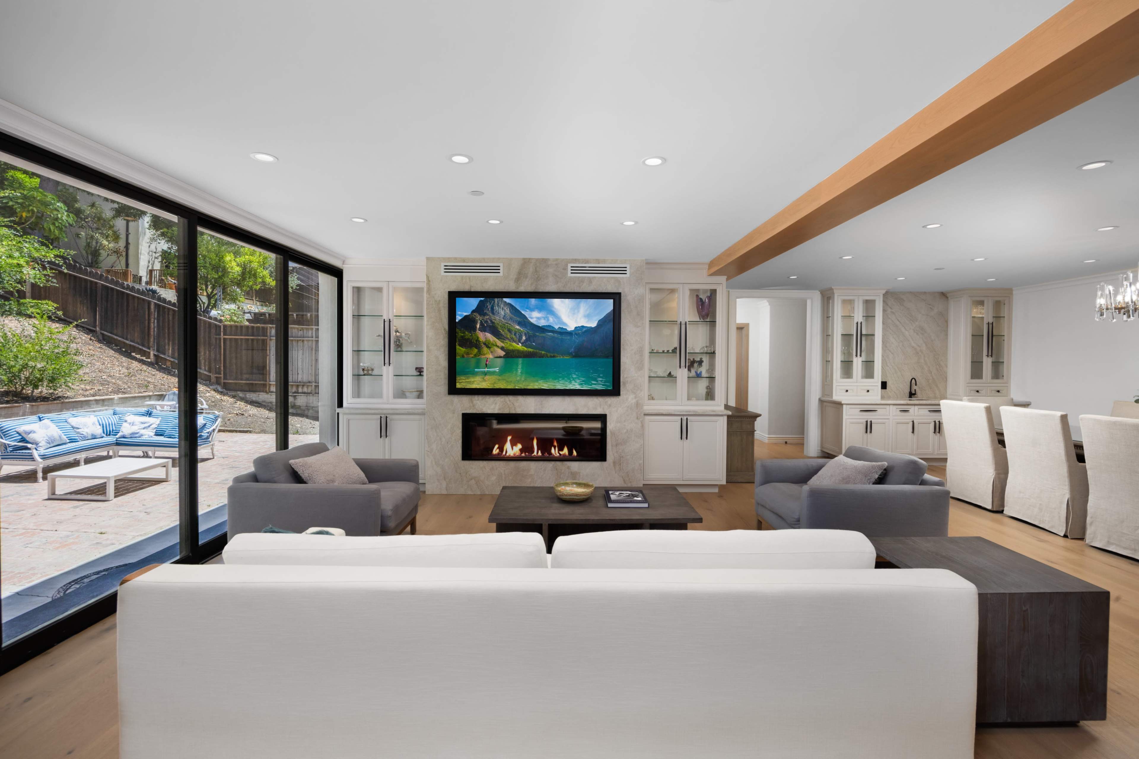 The living room features a large flat-screen TV mounted above a modern fireplace, with seating arranged around a coffee table and large windows providing a view of an outdoor patio.