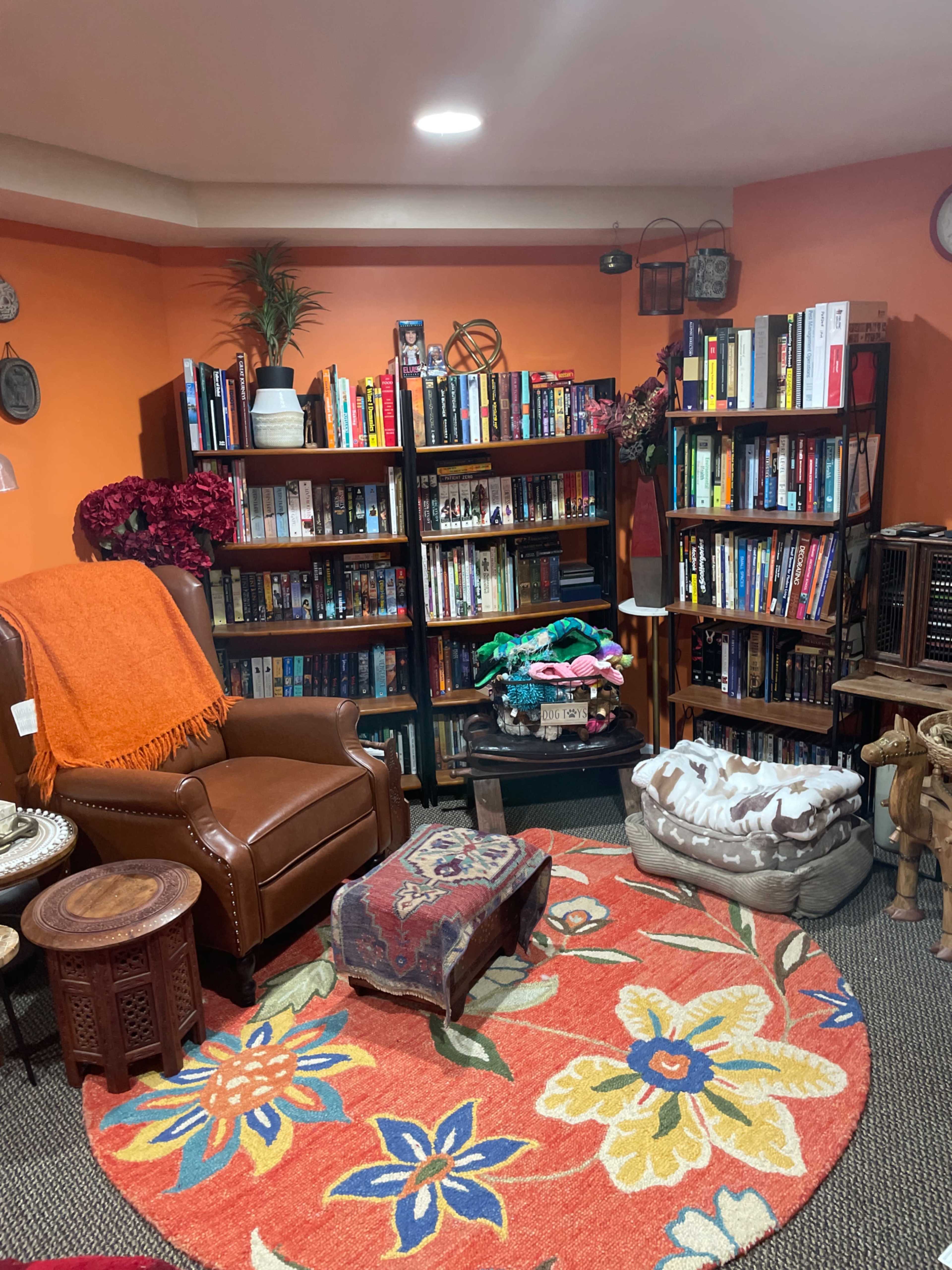 The image shows a cozy room with orange walls, featuring a leather armchair, bookshelves filled with a variety of books, and a colorful area rug with floral patterns.