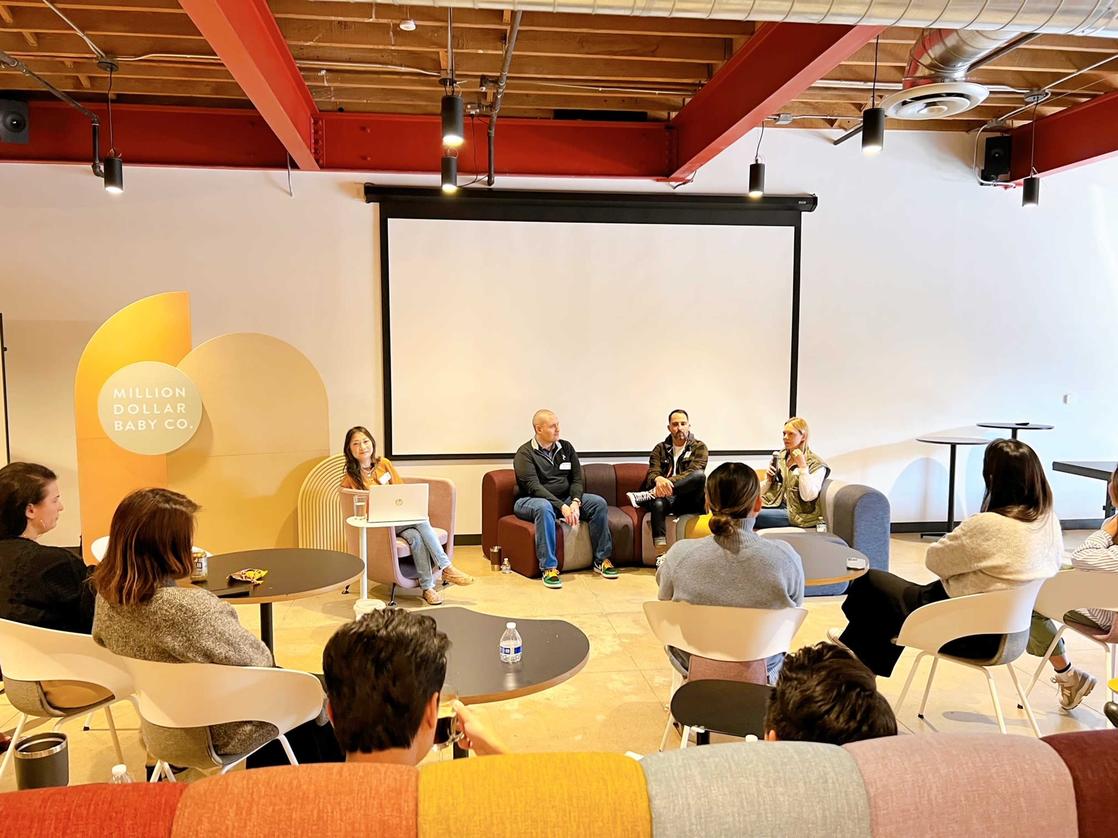 A panel discussion is taking place in a modern conference room, with a seated audience facing four speakers at the front.