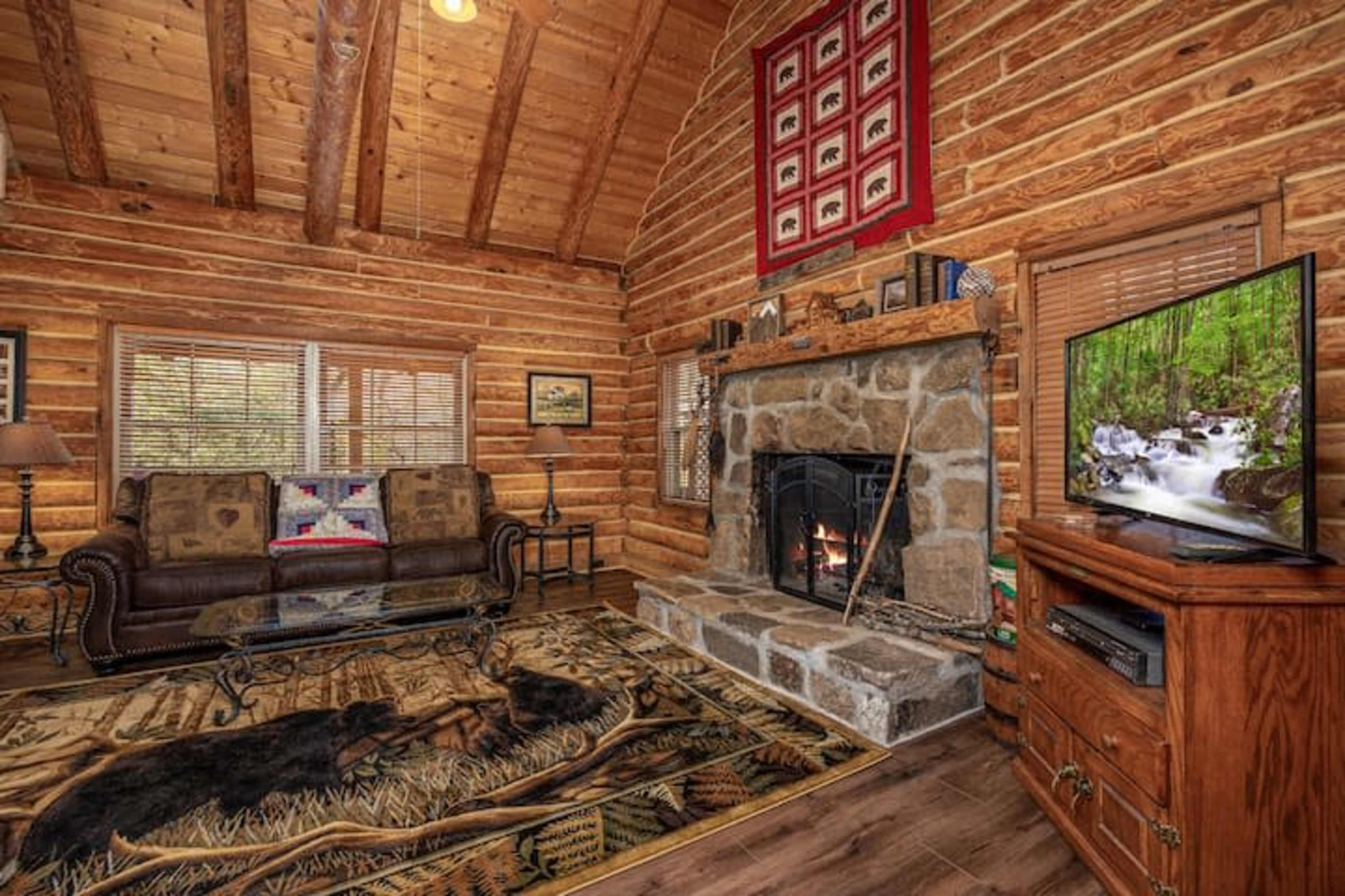 The interior of a log cabin features a stone fireplace, wooden beams, a couch, and a television, with a decorative rug on the floor and artwork on the walls.