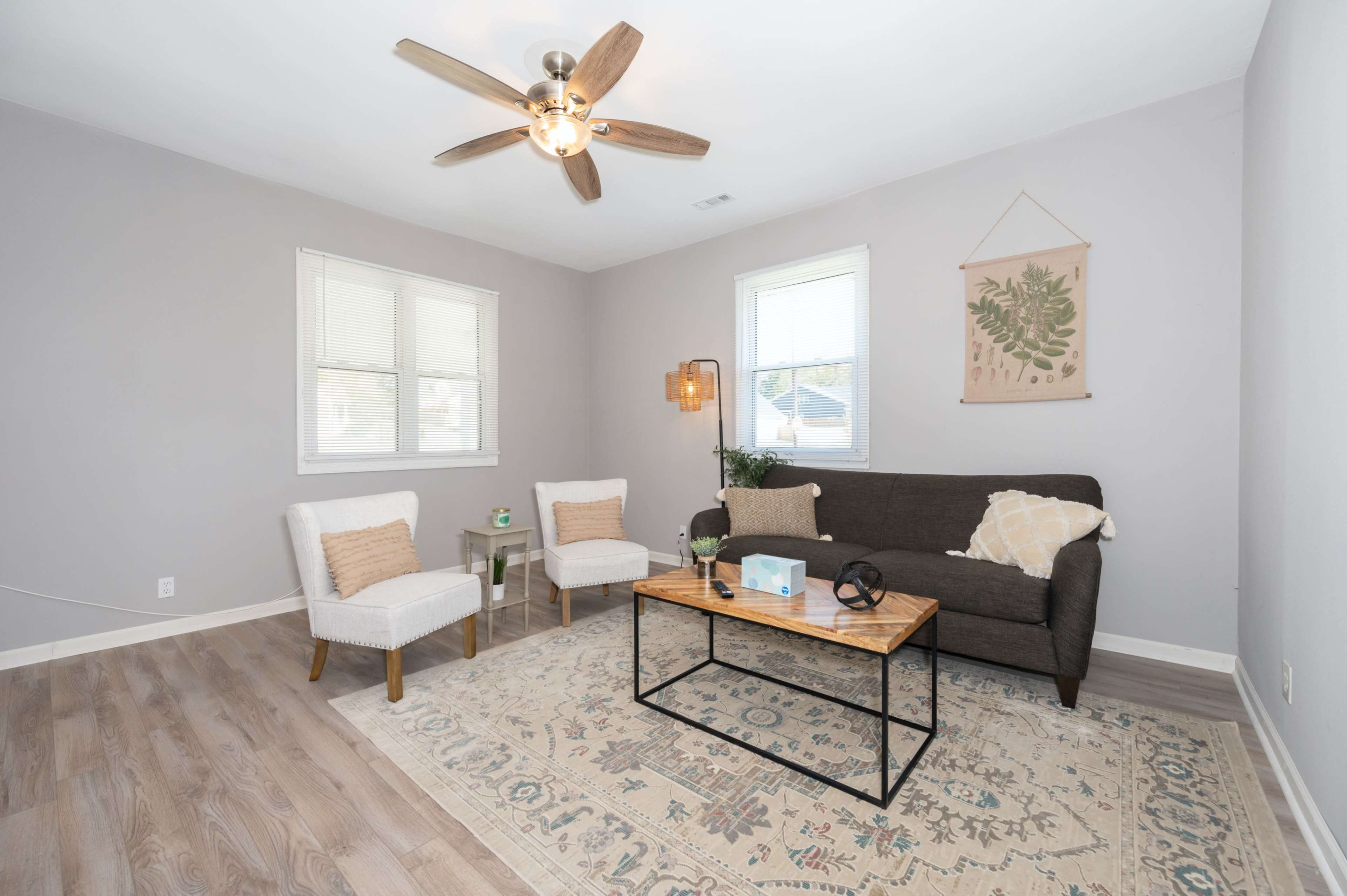 The living room features a brown sofa, two white chairs, a coffee table, and a ceiling fan, all arranged on a wooden floor with a decorative area rug.