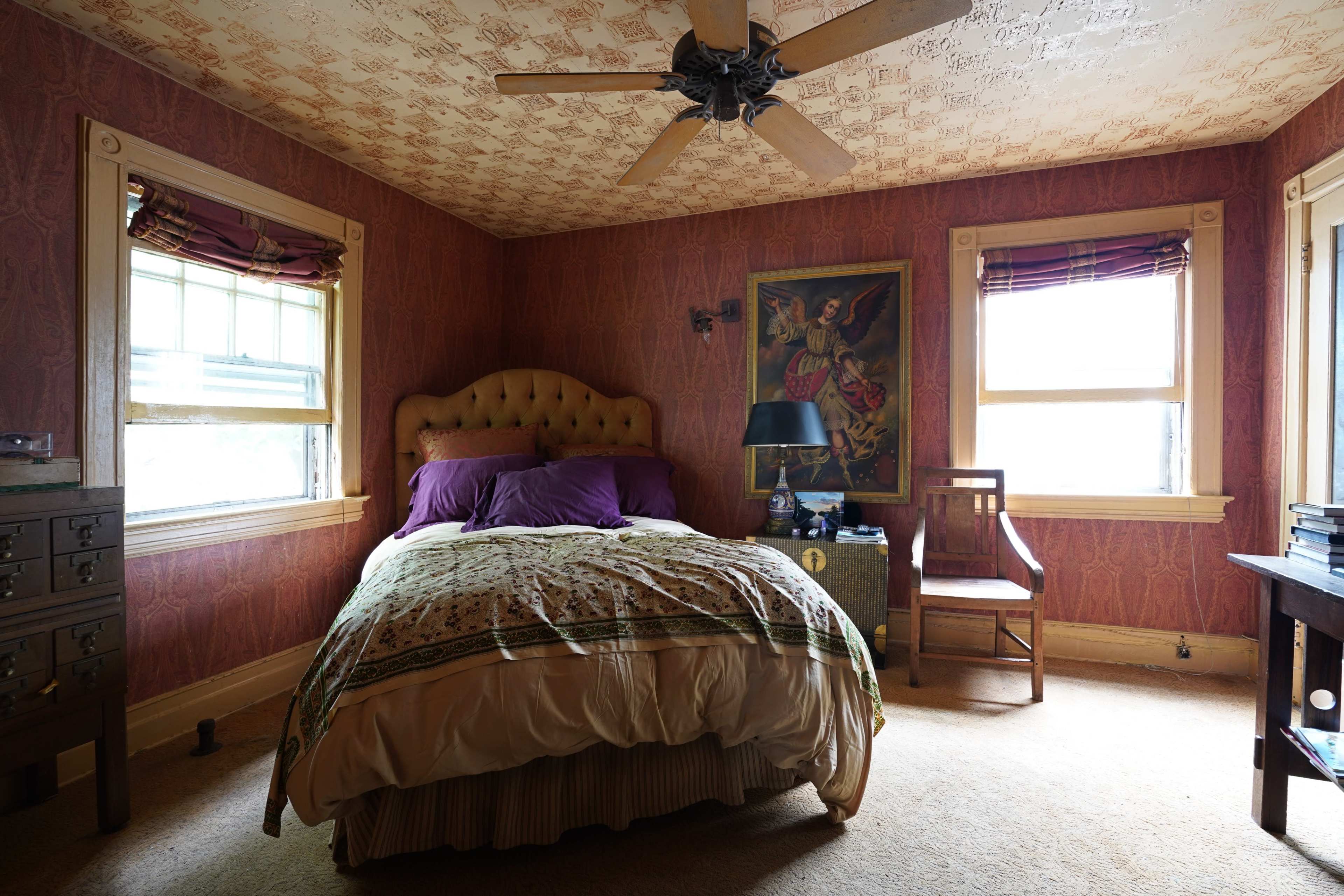 A bedroom featuring a large bed with a patterned comforter, two windows, a wooden chair, and a painting on the wall.