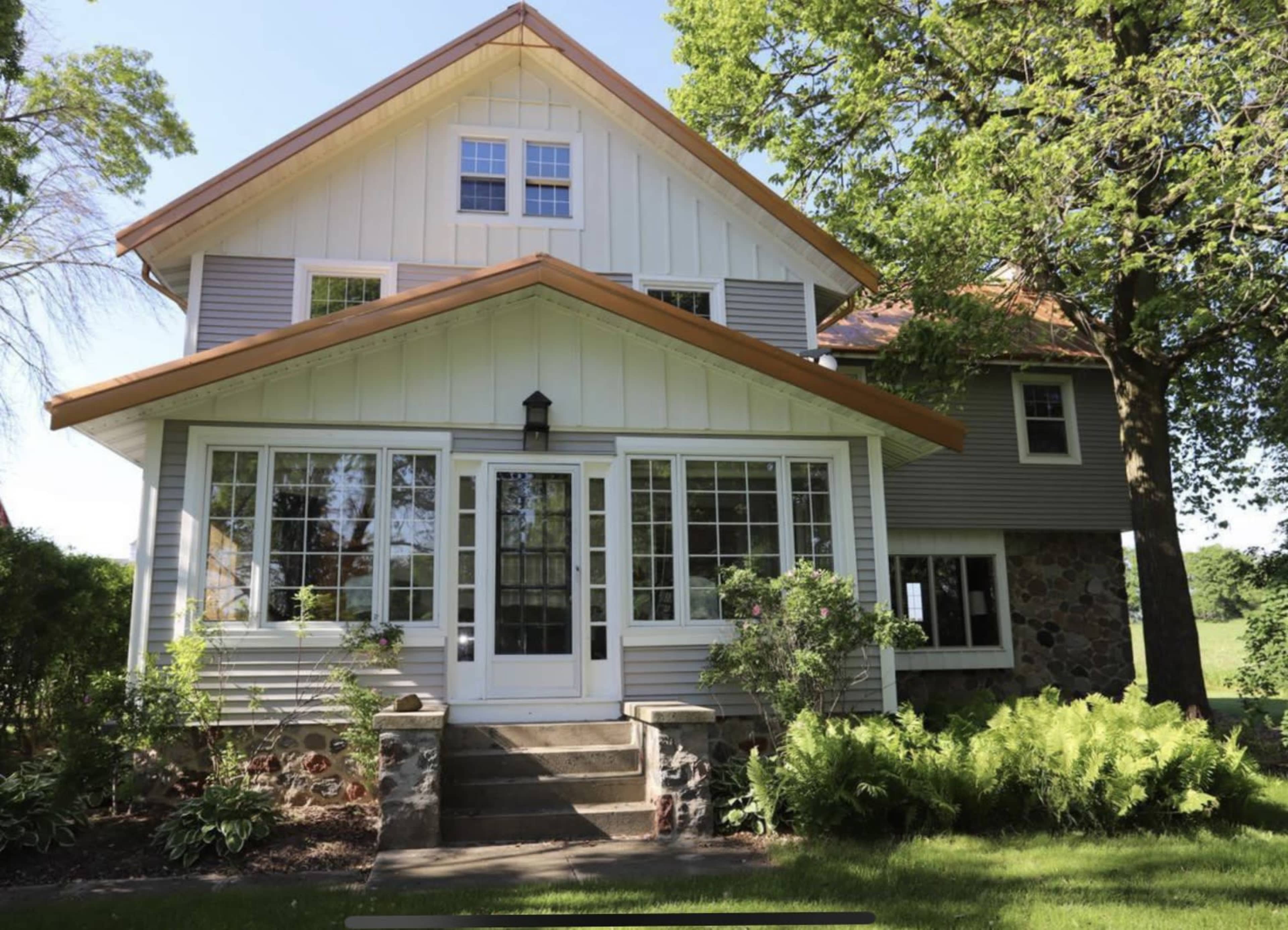 A two-story house features a front porch with large windows, surrounded by greenery and a stone foundation.
