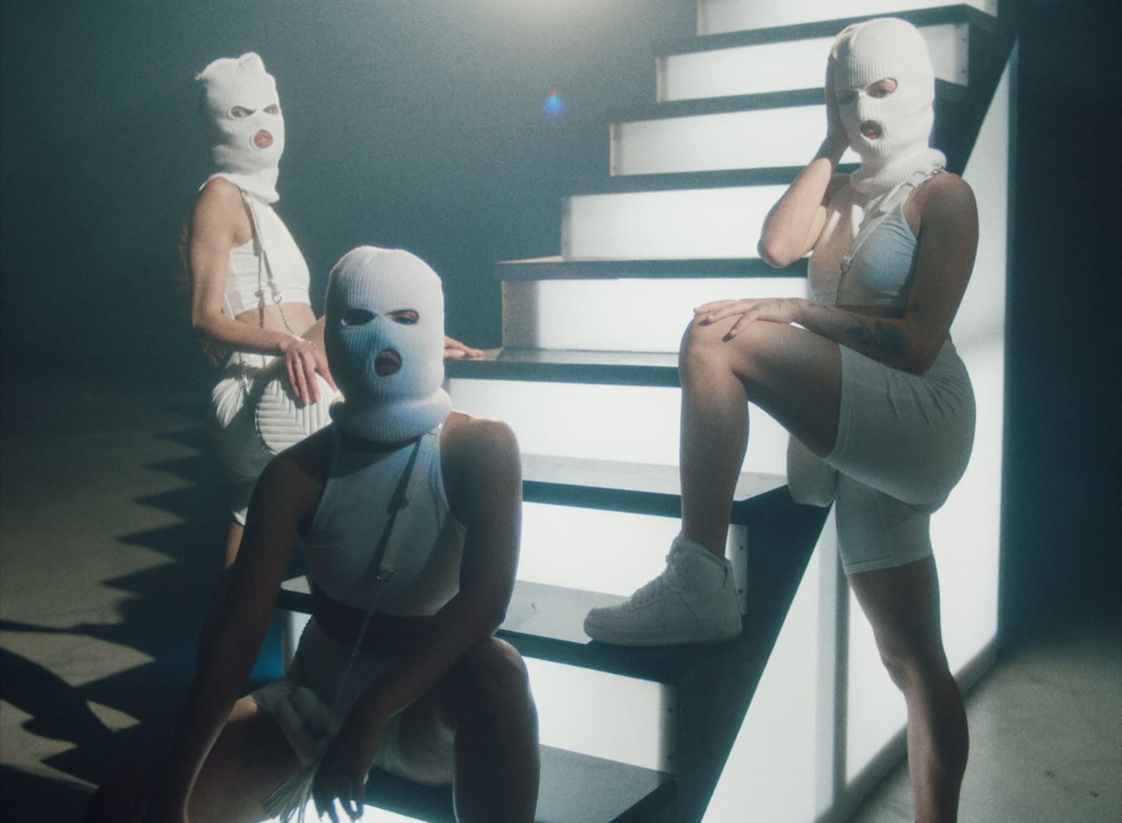 Three individuals wearing white balaclavas and matching outfits pose on a set of illuminated stairs in a dimly lit environment.