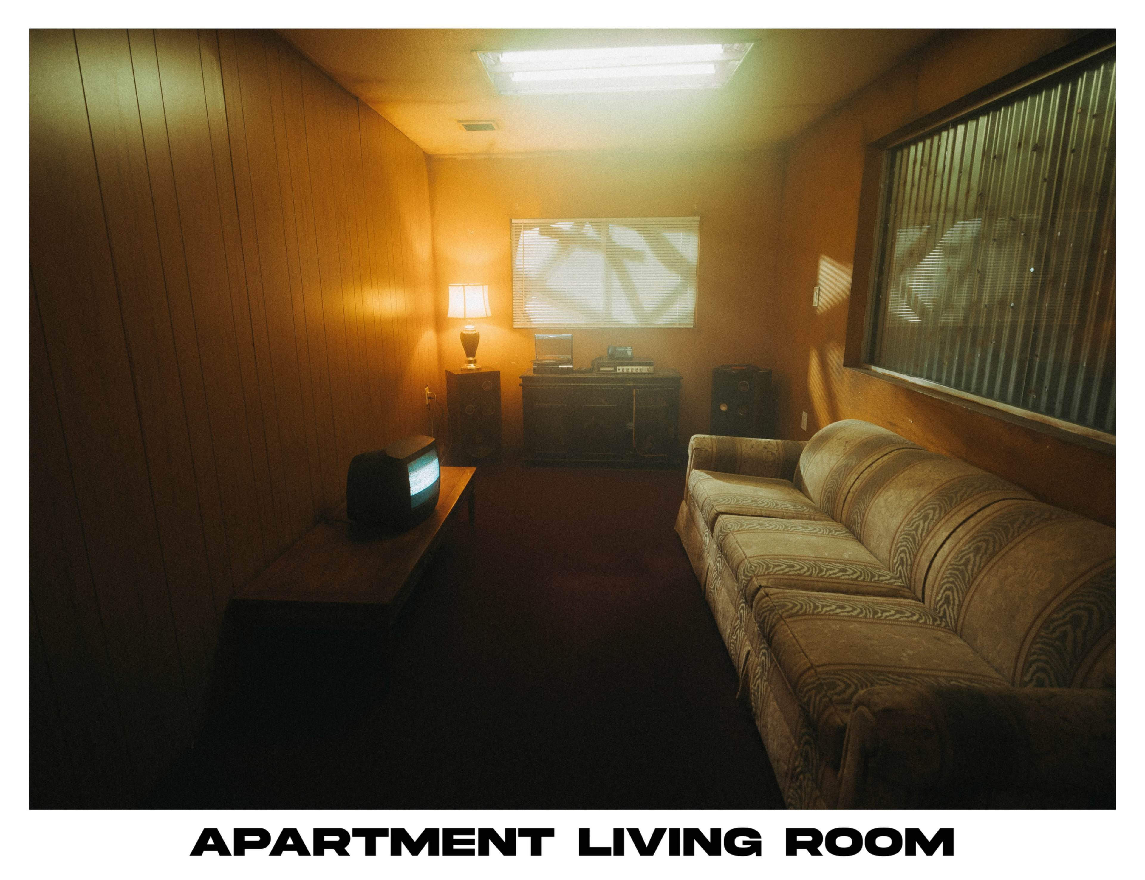 A dimly lit apartment living room with a patterned couch, a small table lamp, an old television, and a wooden cabinet.