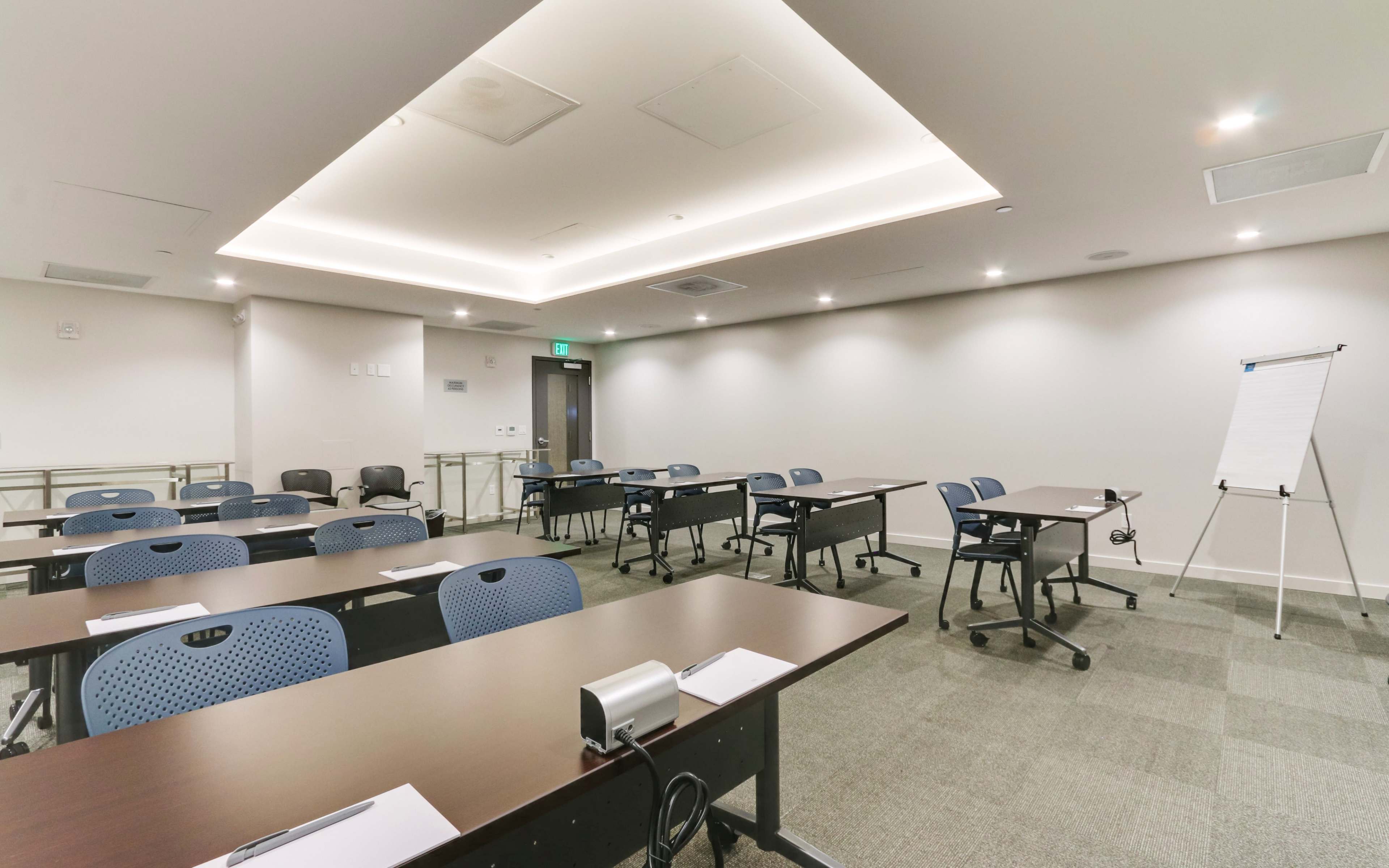 A well-lit meeting room features multiple rows of desks and chairs with a flip chart at one end.