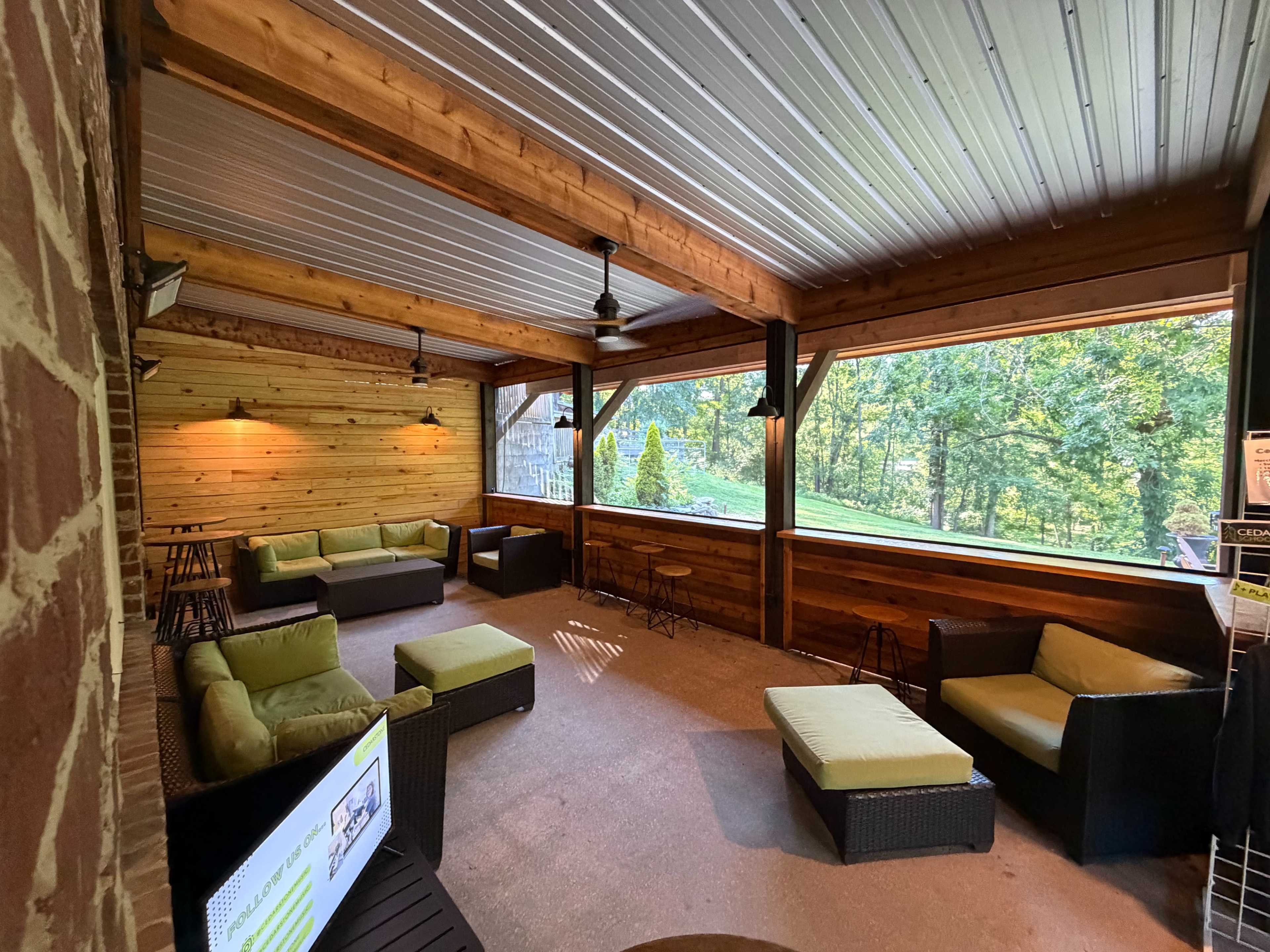 The image shows a screened-in porch featuring several seating arrangements with green cushions and wooden walls, overlooking a forested area.