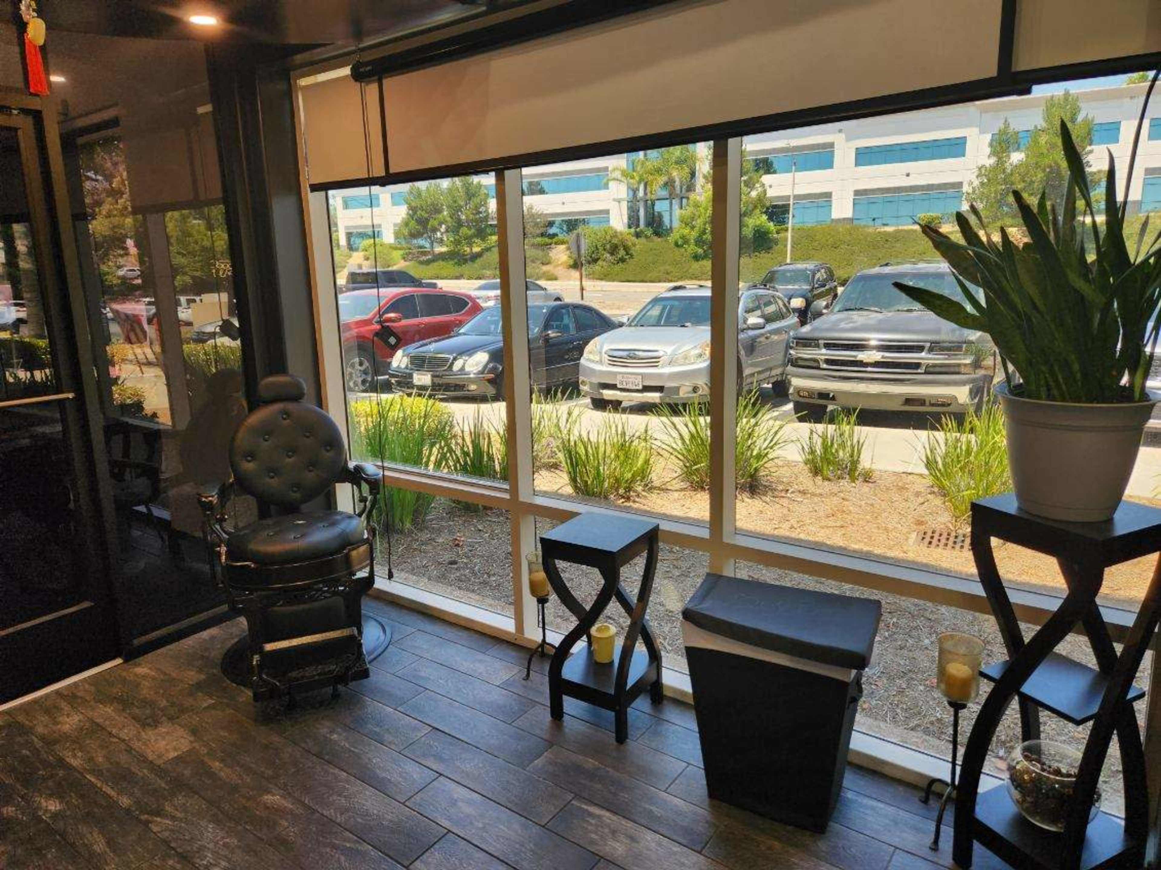 The image shows a barber shop interior with a black barber chair next to large windows overlooking parked cars and greenery outside.