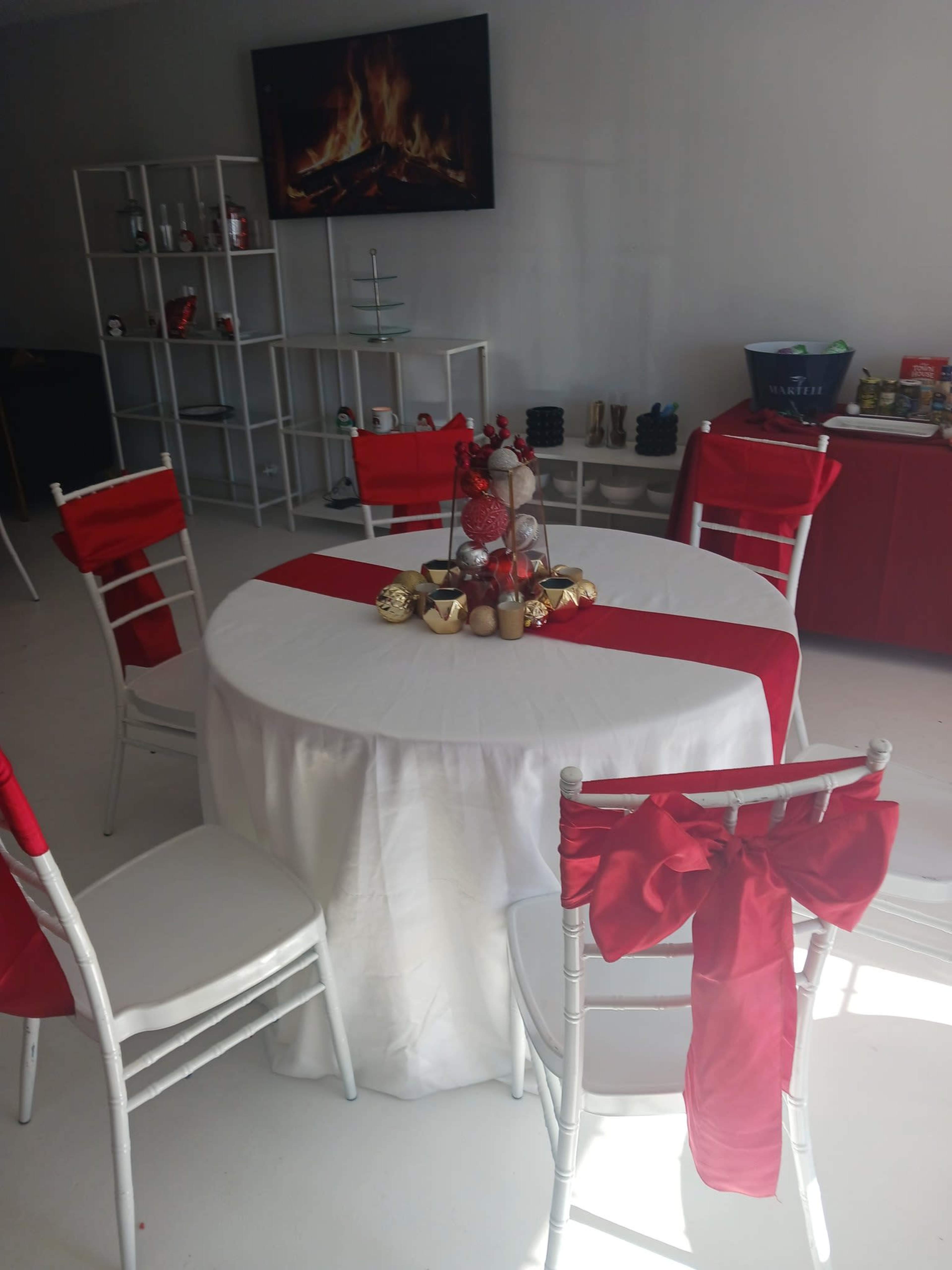 A round table draped in a white tablecloth with a red runner is set in a room with several chairs, a decorative centerpiece, and a wall-mounted television displaying a fireplace image.