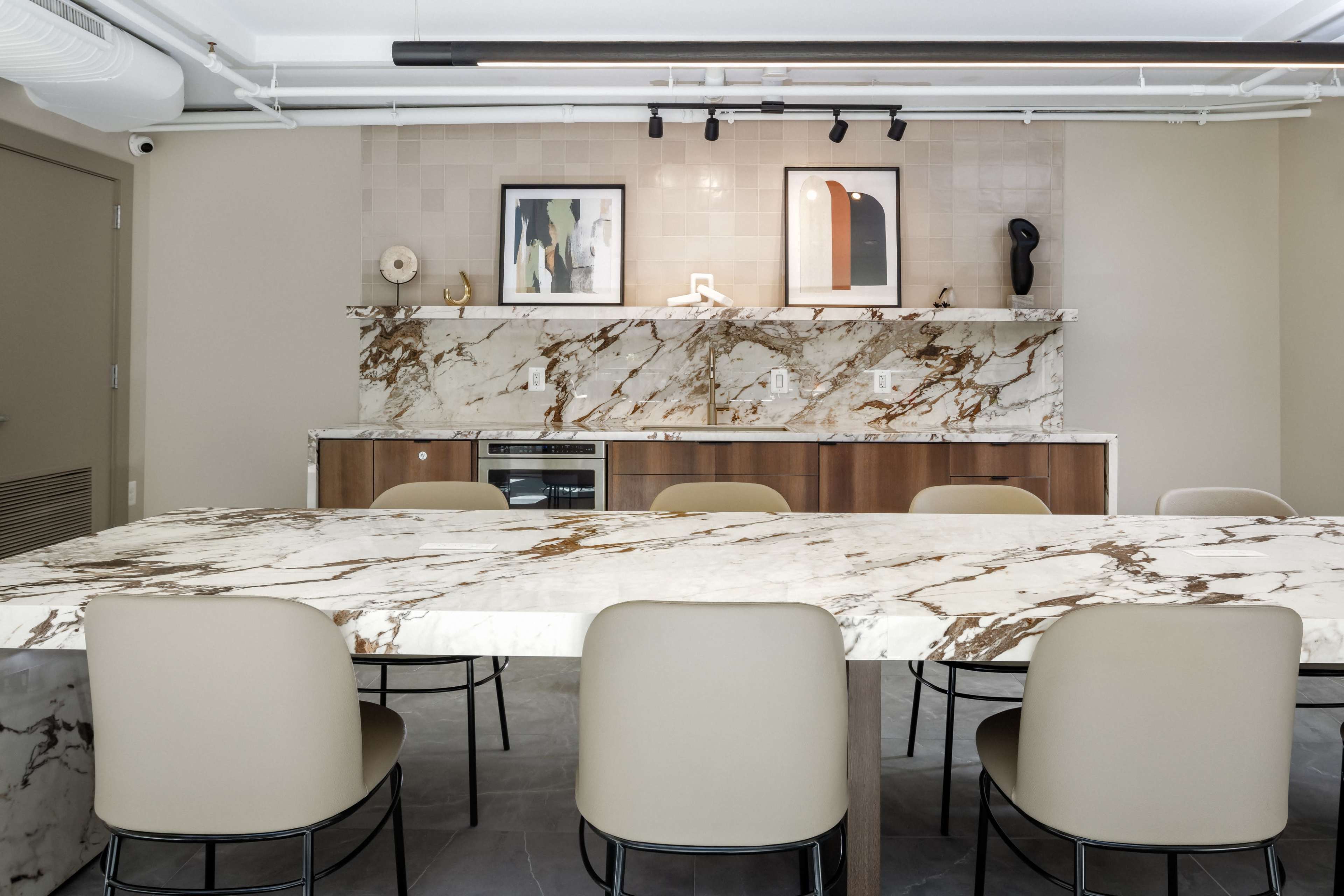 A modern kitchen features a large marble countertop with beige chairs and a stone backsplash displaying framed artwork.