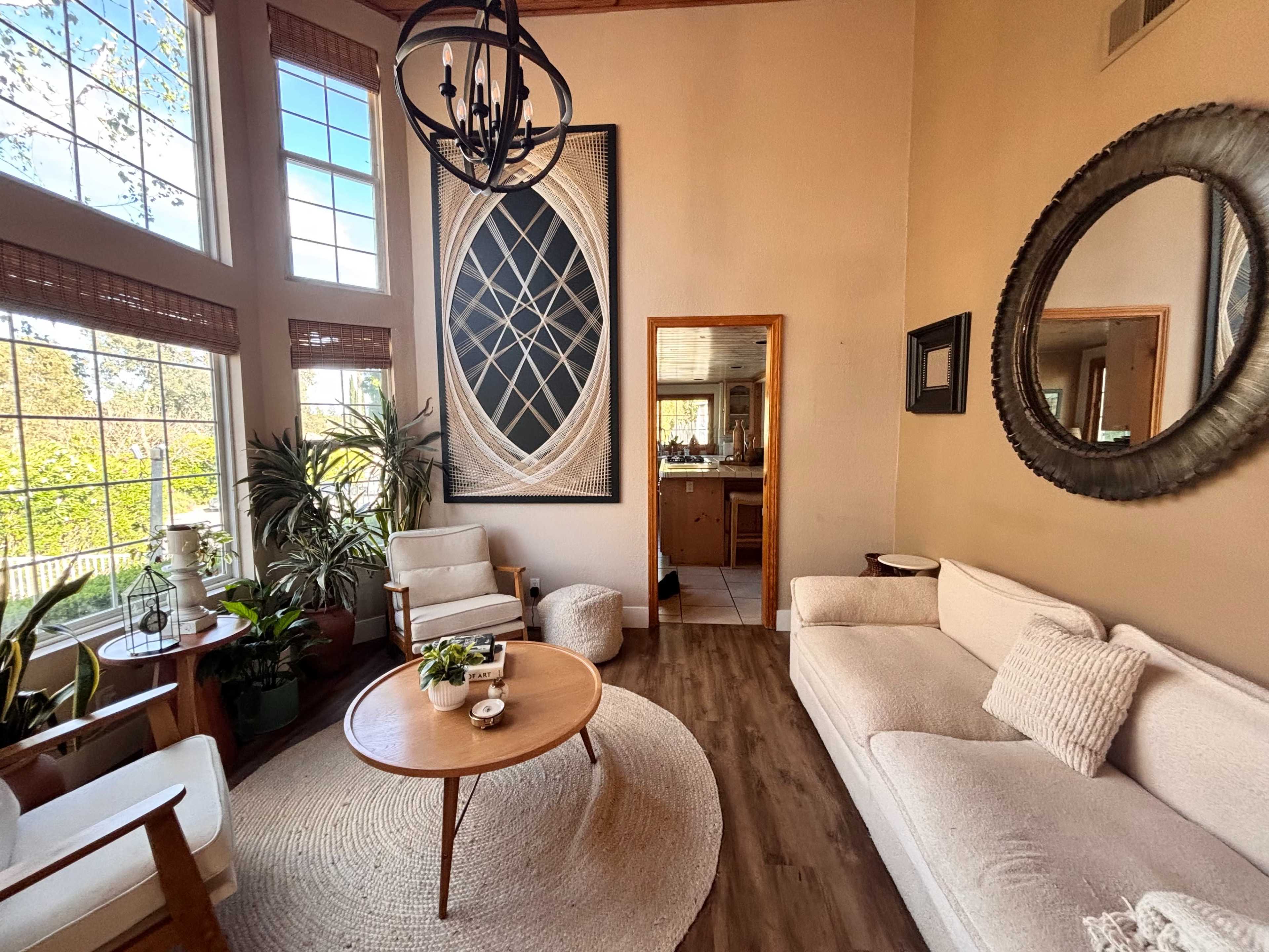 A bright, airy living room features large windows, a round coffee table, a white sofa, and potted plants.