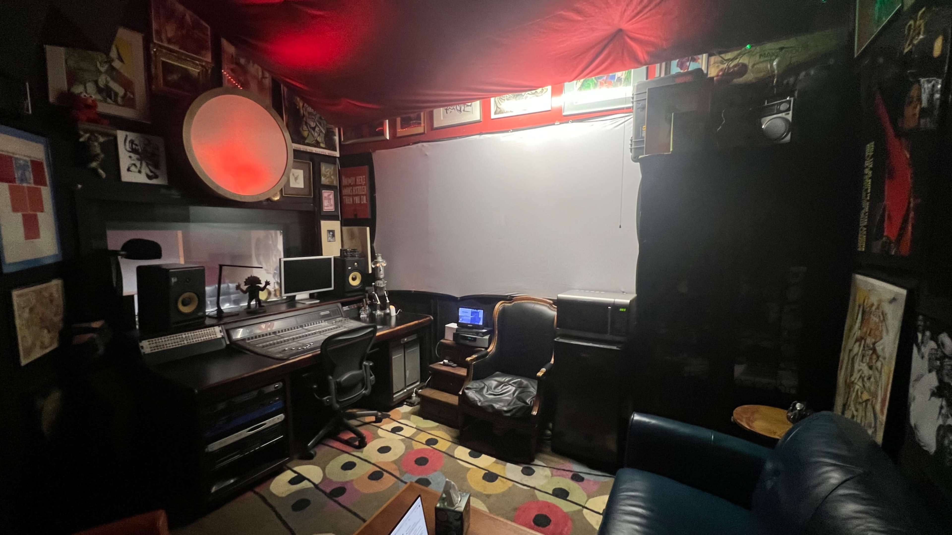 A dimly lit music studio with a mixing console, computer setup, an armchair, and various framed artworks on the walls.