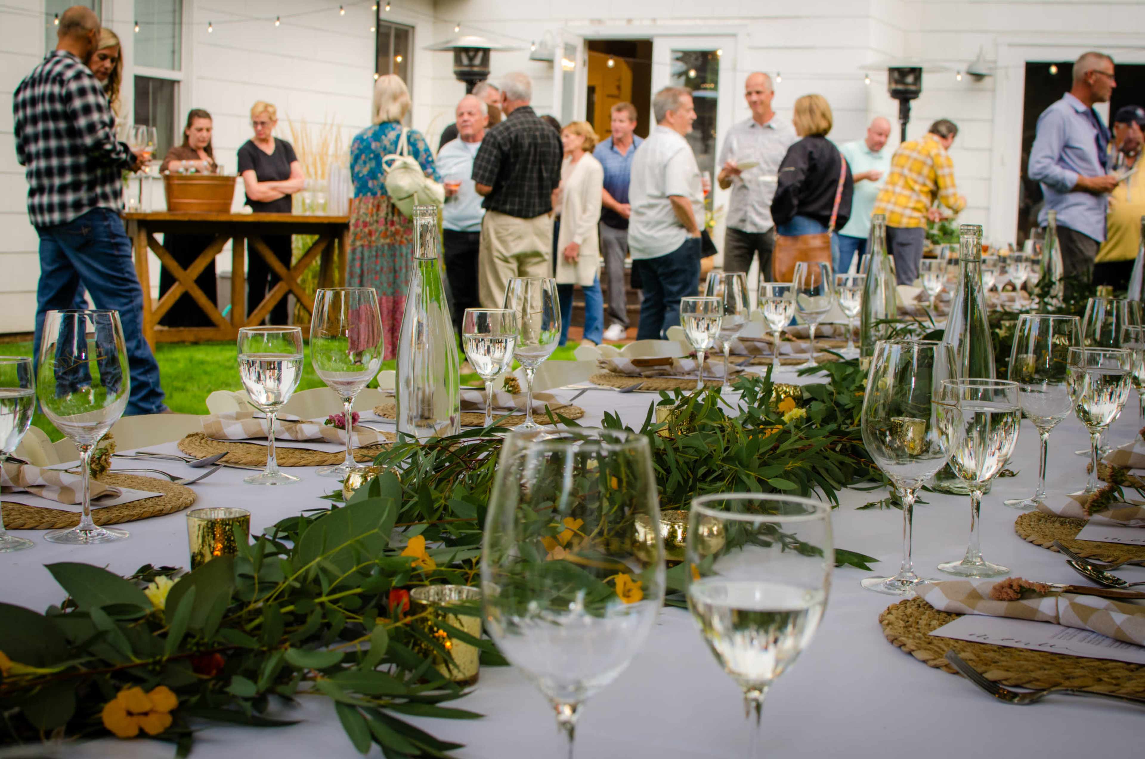 A table is set for a gathering with glasses and greenery, while guests socialize in the background at an outdoor event.