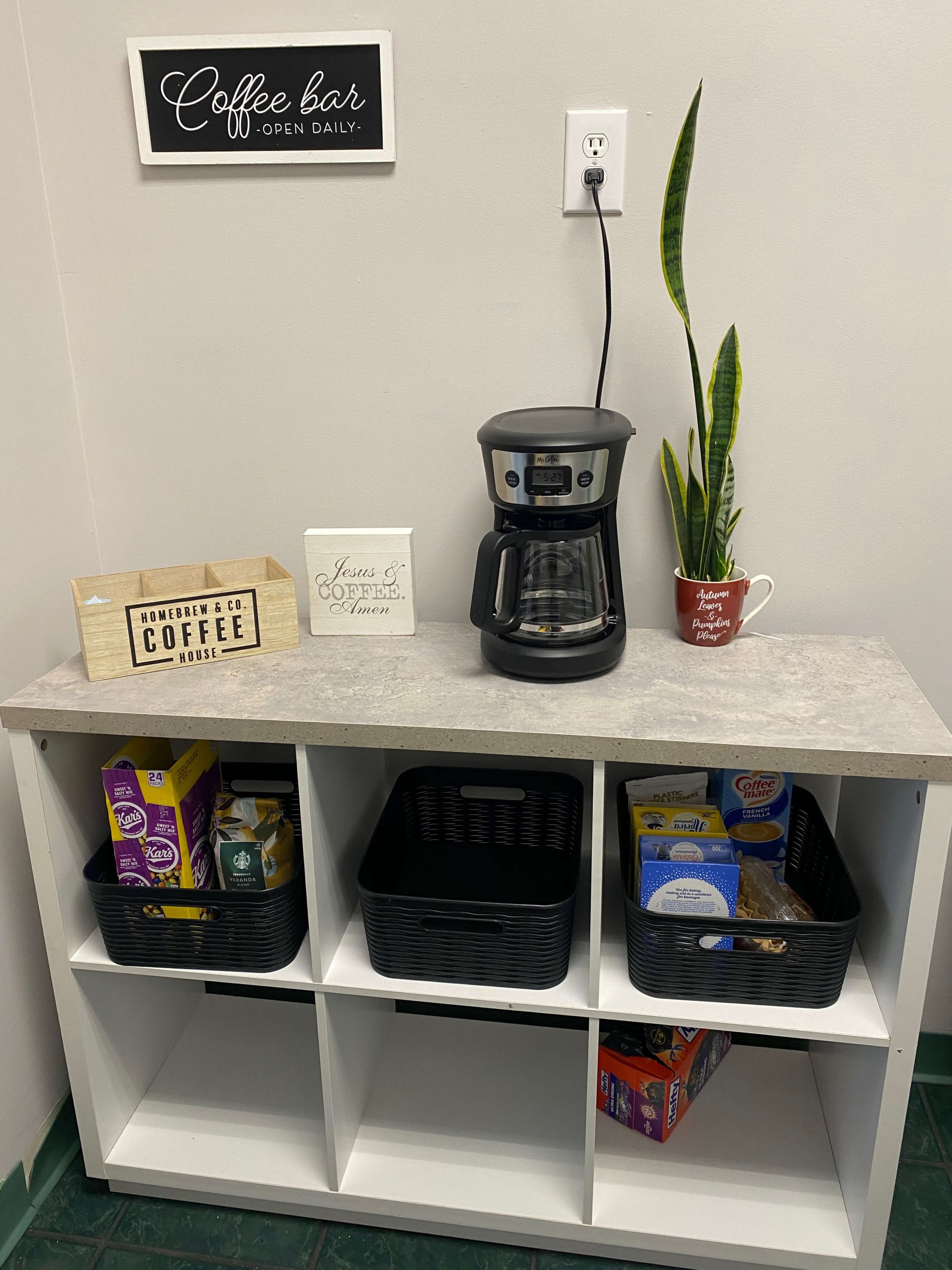 The image shows a small coffee station featuring a coffee maker, decorative signs, and organized storage baskets on a white shelf.