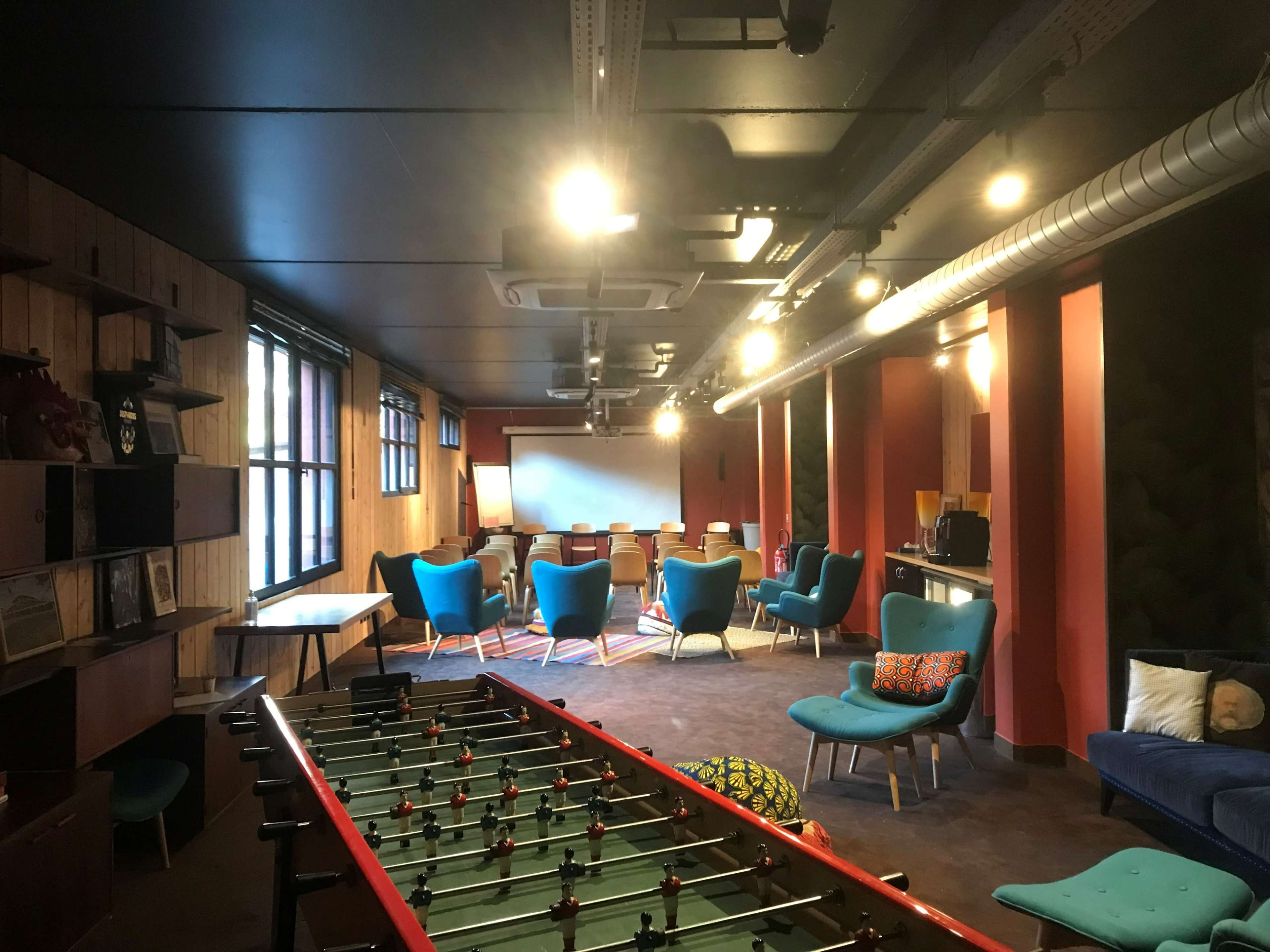 A modern recreational room with a foosball table in the foreground and a seating area featuring blue chairs and a projector screen at the back.