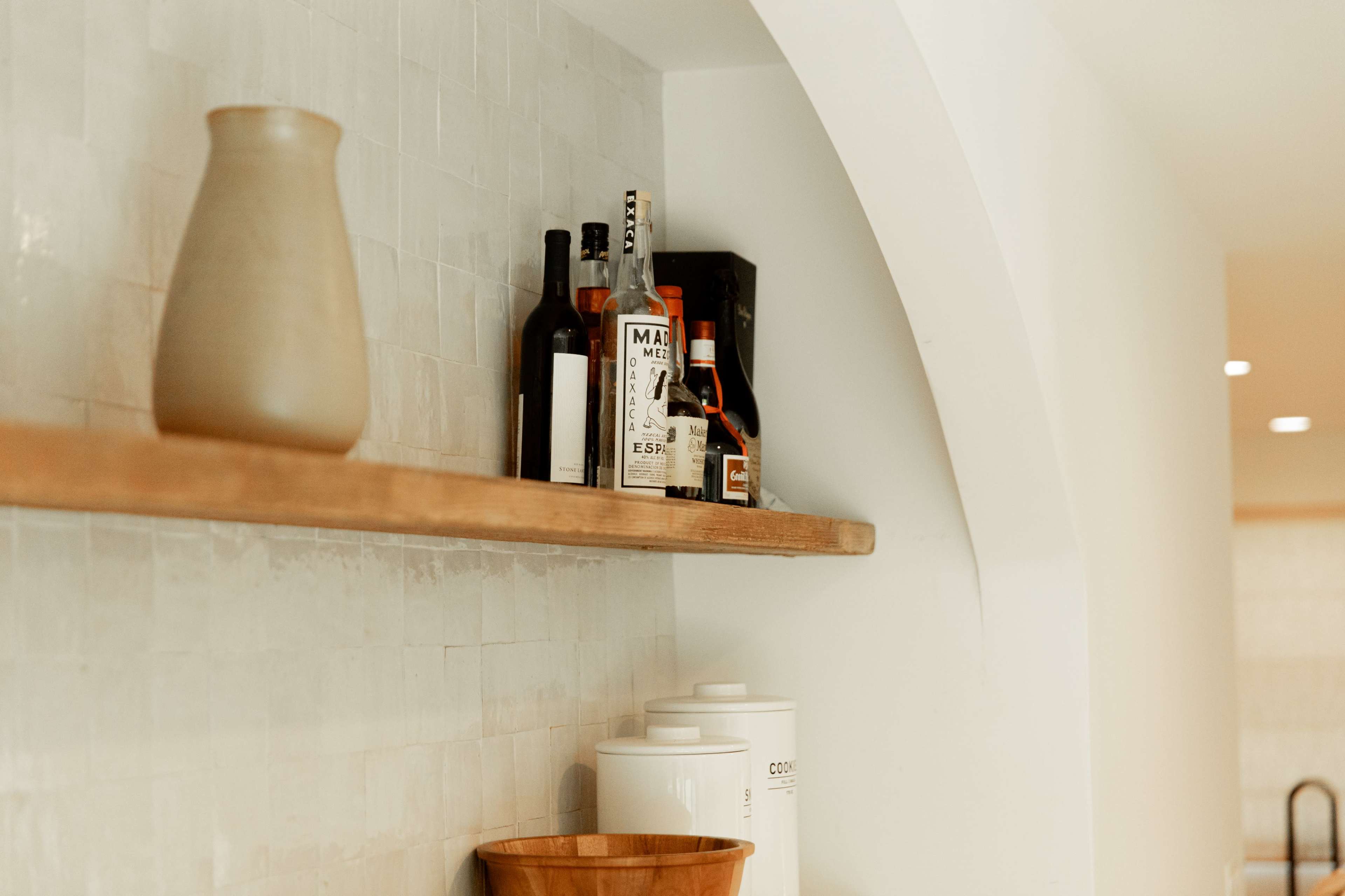 A wooden shelf mounted on a white wall holds several bottles of liquor and a ceramic pitcher.