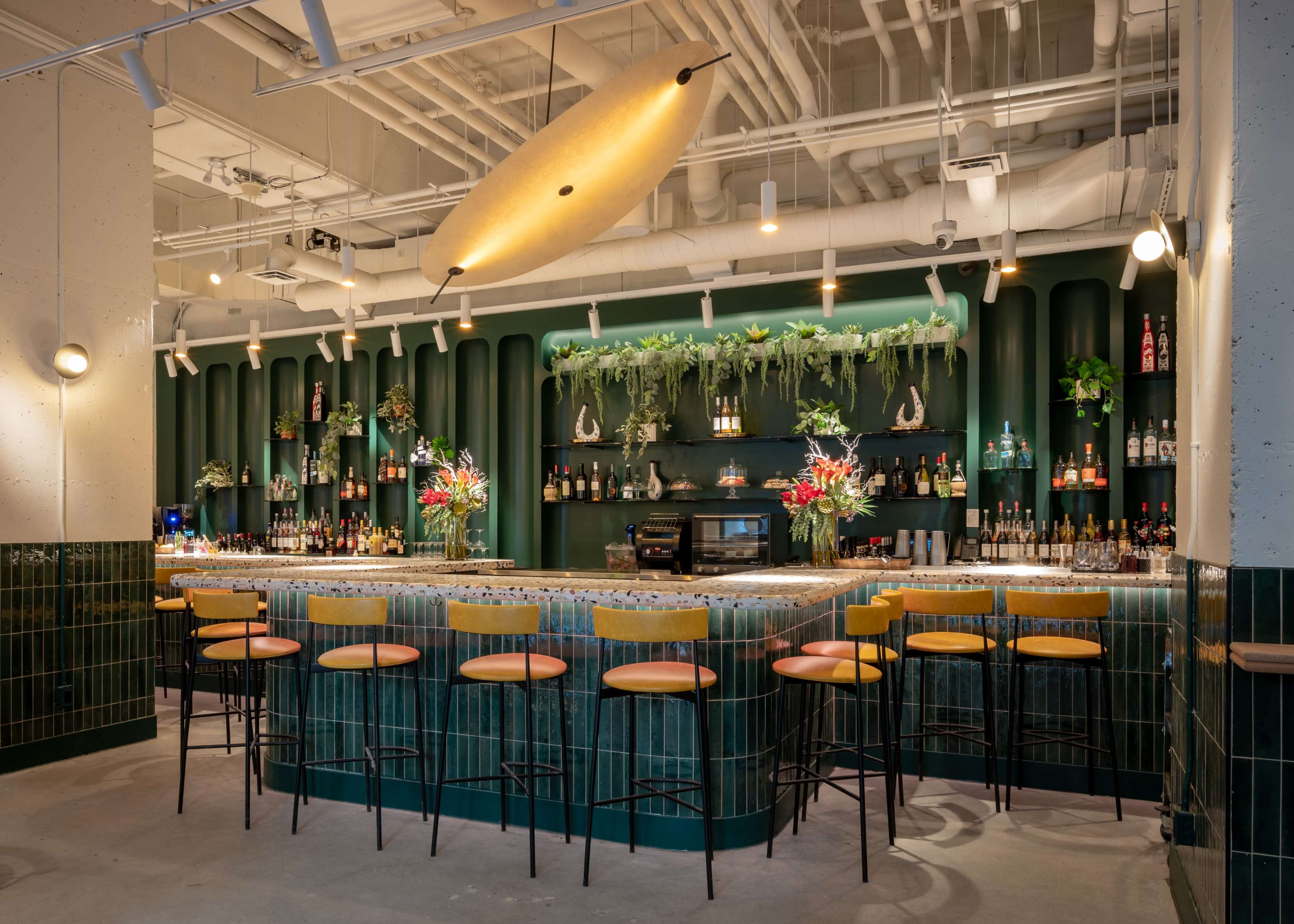 The image shows a modern bar with a green and gold color scheme, featuring a curved counter, high stools, and shelves stocked with various bottles, adorned with plants and decorative elements.