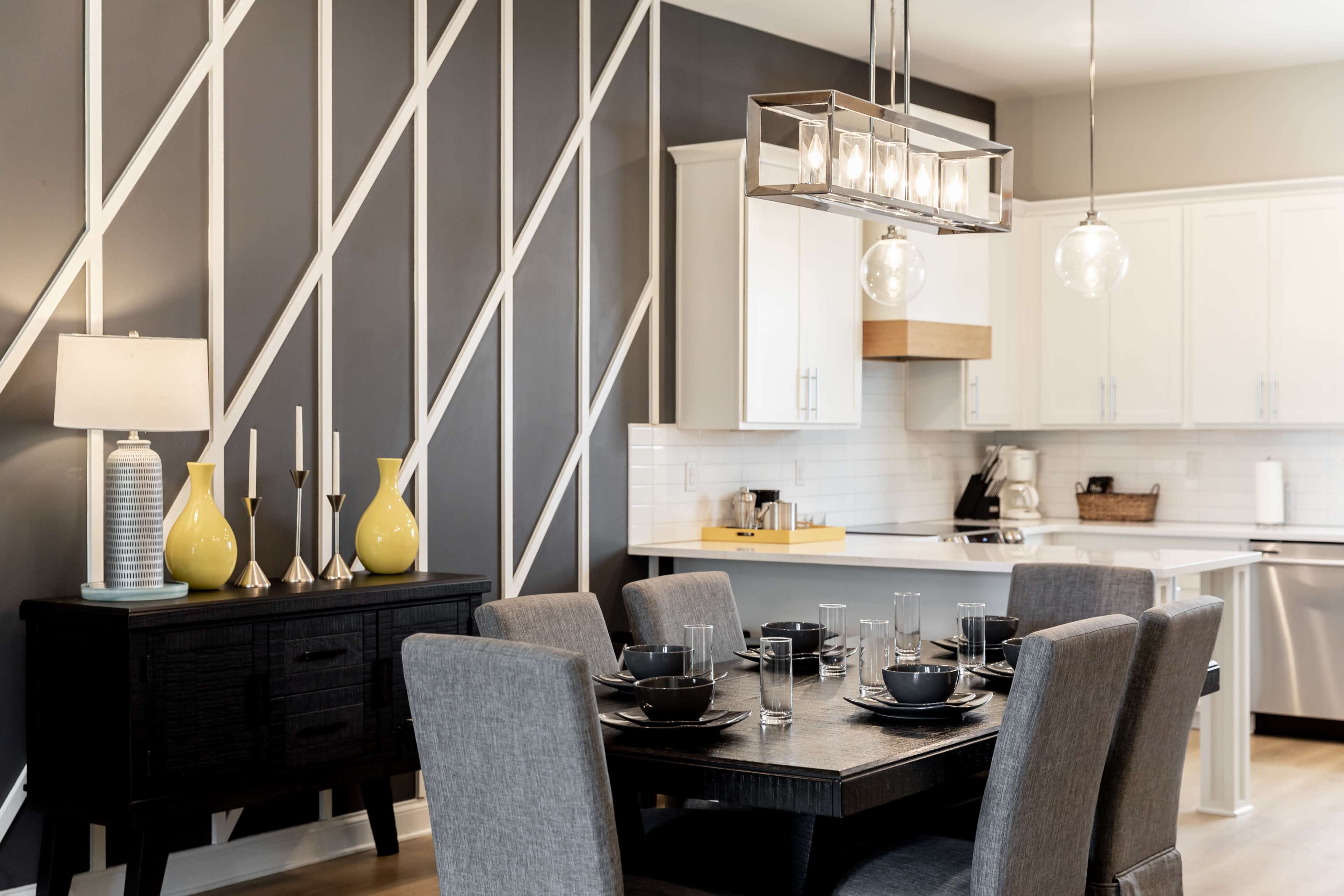 The image shows a modern dining area with a black table set for six, surrounded by gray chairs, and featuring a kitchen in the background with white cabinetry and a decorative wall.