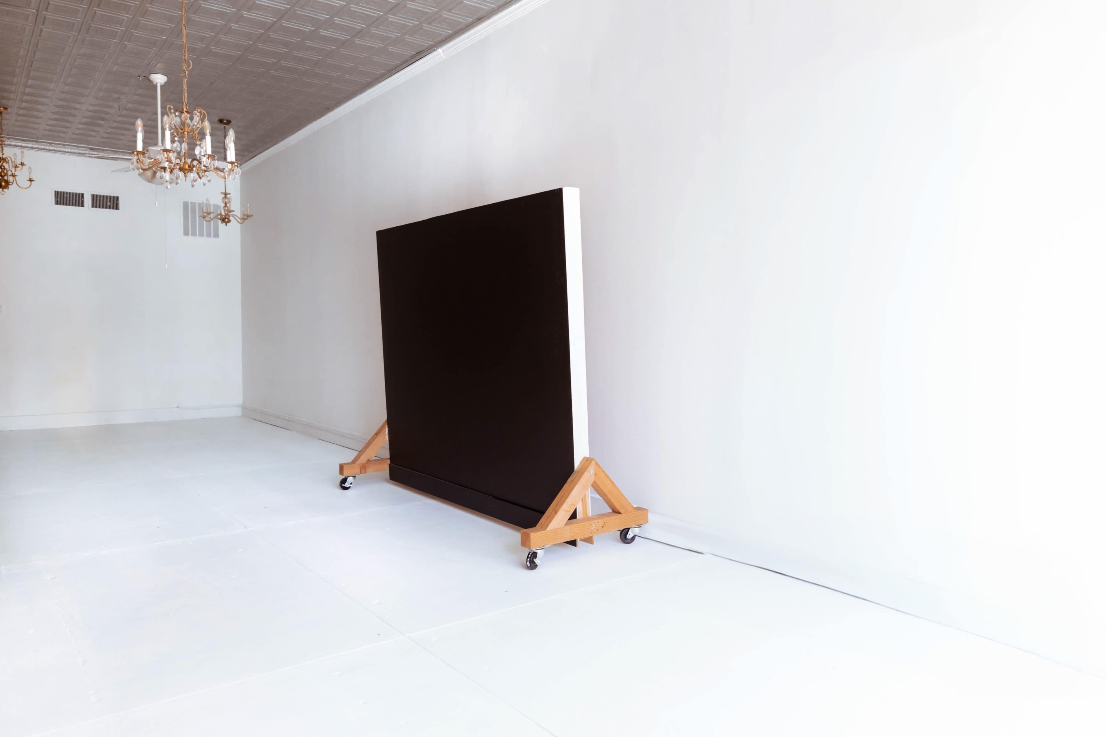 The image shows a large black canvas on a wooden easel with wheels, positioned against a plain white wall in a minimalistic room.