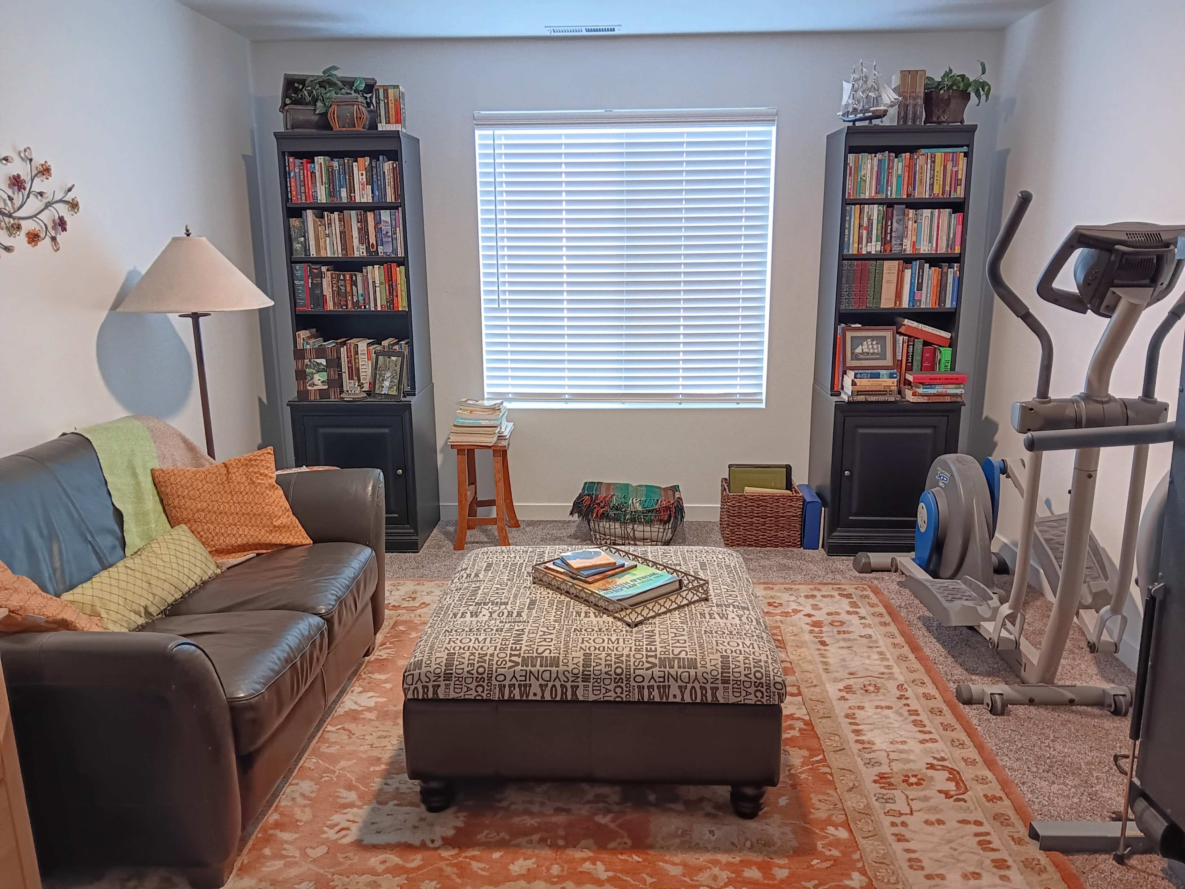 A cozy room features a leather sofa, an ottoman, two tall bookshelves filled with books, and an exercise machine beside a window with blinds.