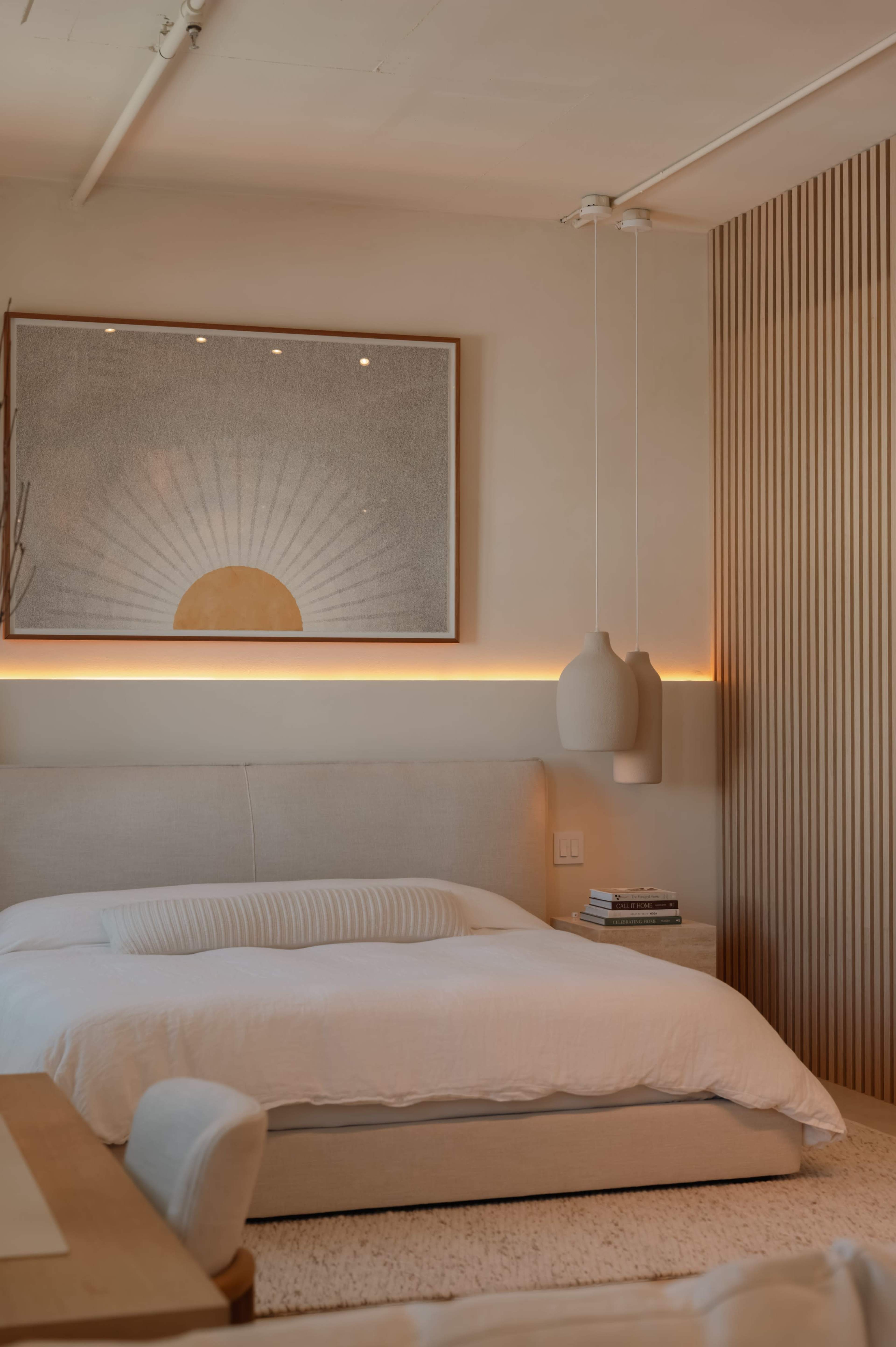 The image shows a modern bedroom with a neutral color palette, featuring a bed with a white duvet, a framed artwork above the headboard, and warm lighting along the wall.