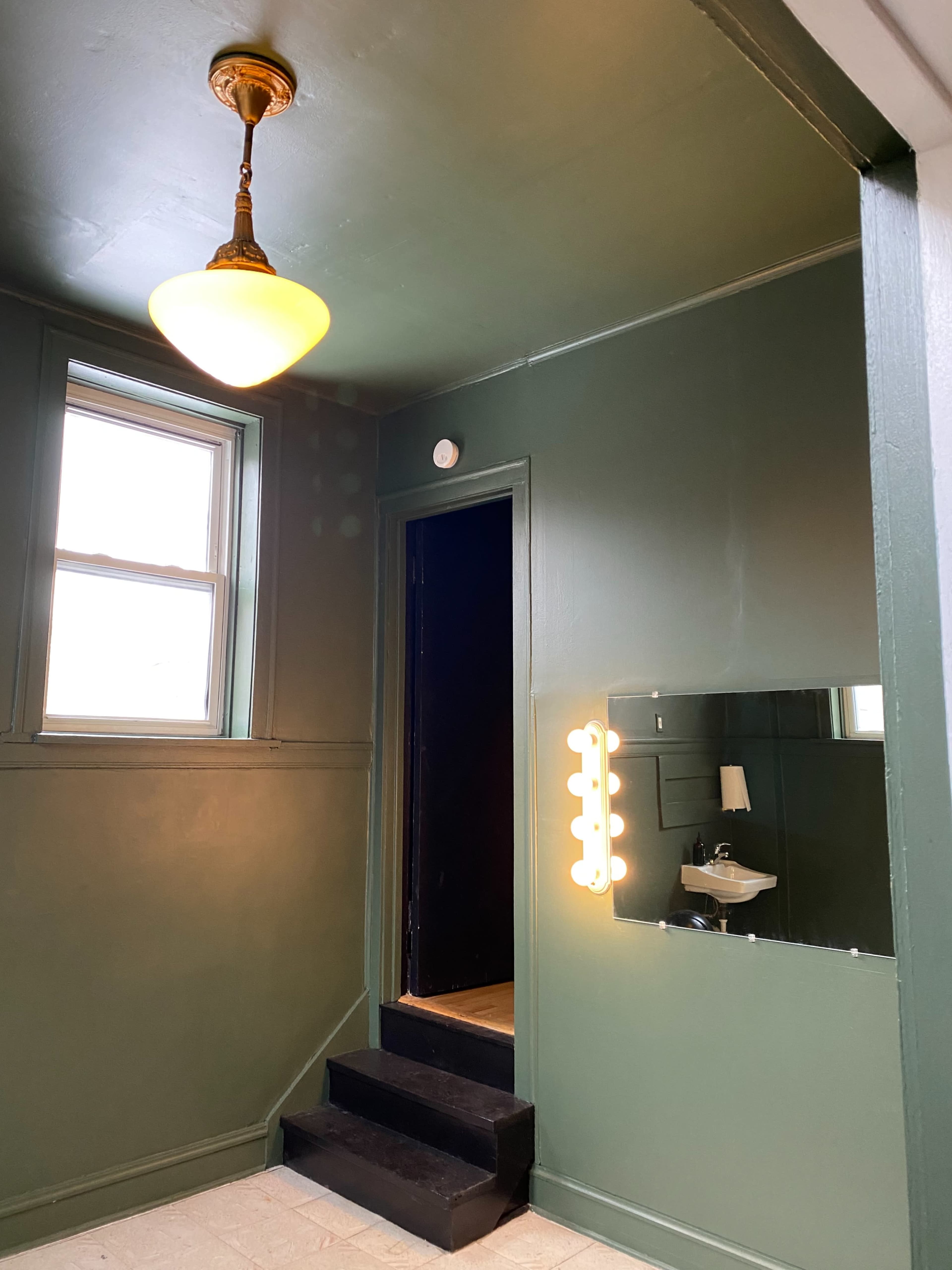 A dimly lit green-walled room with a staircase, a small sink, and a mirror framed by bright lights.