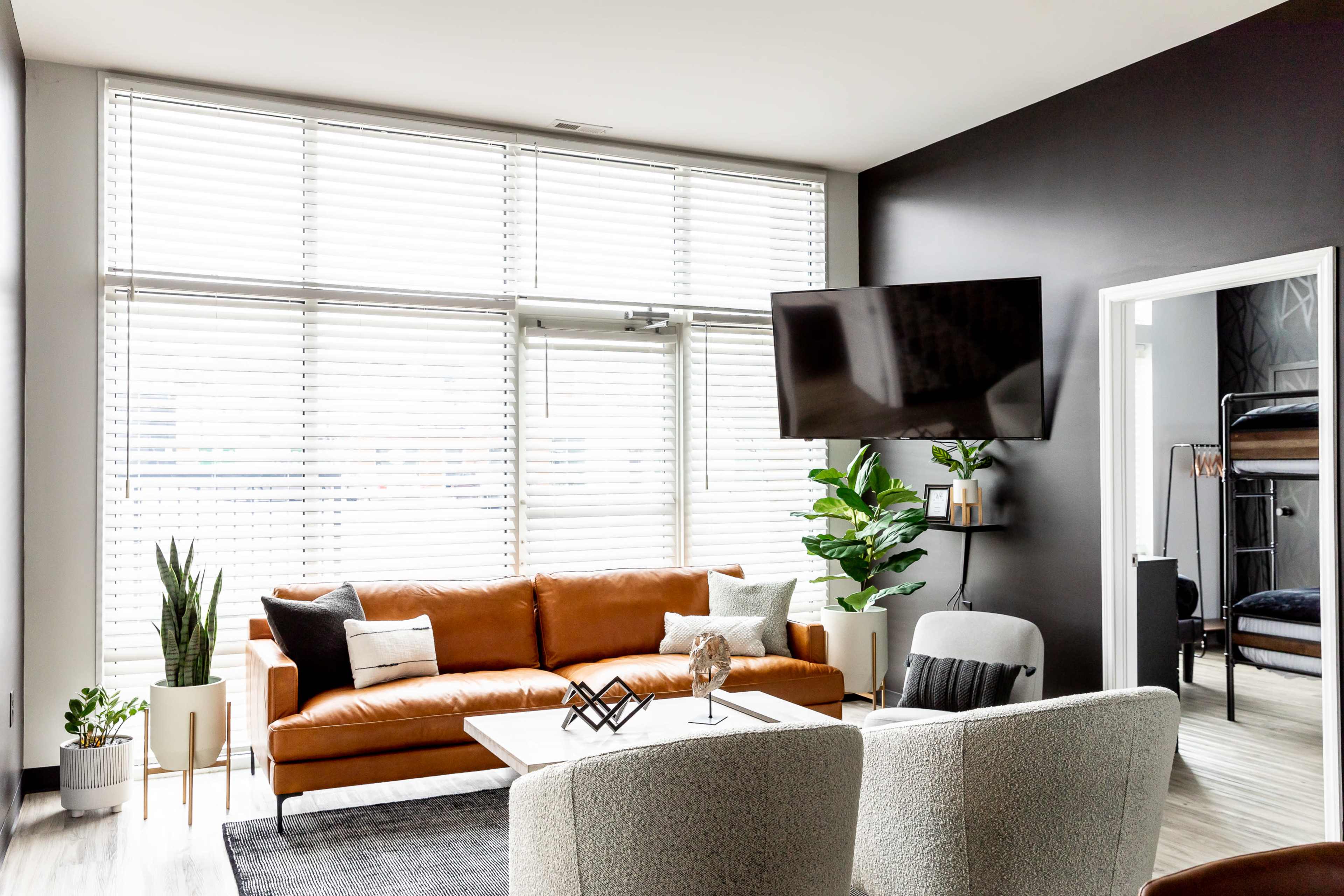 The living room features a brown leather sofa, a coffee table, large windows with blinds, and a mounted television against a dark wall.