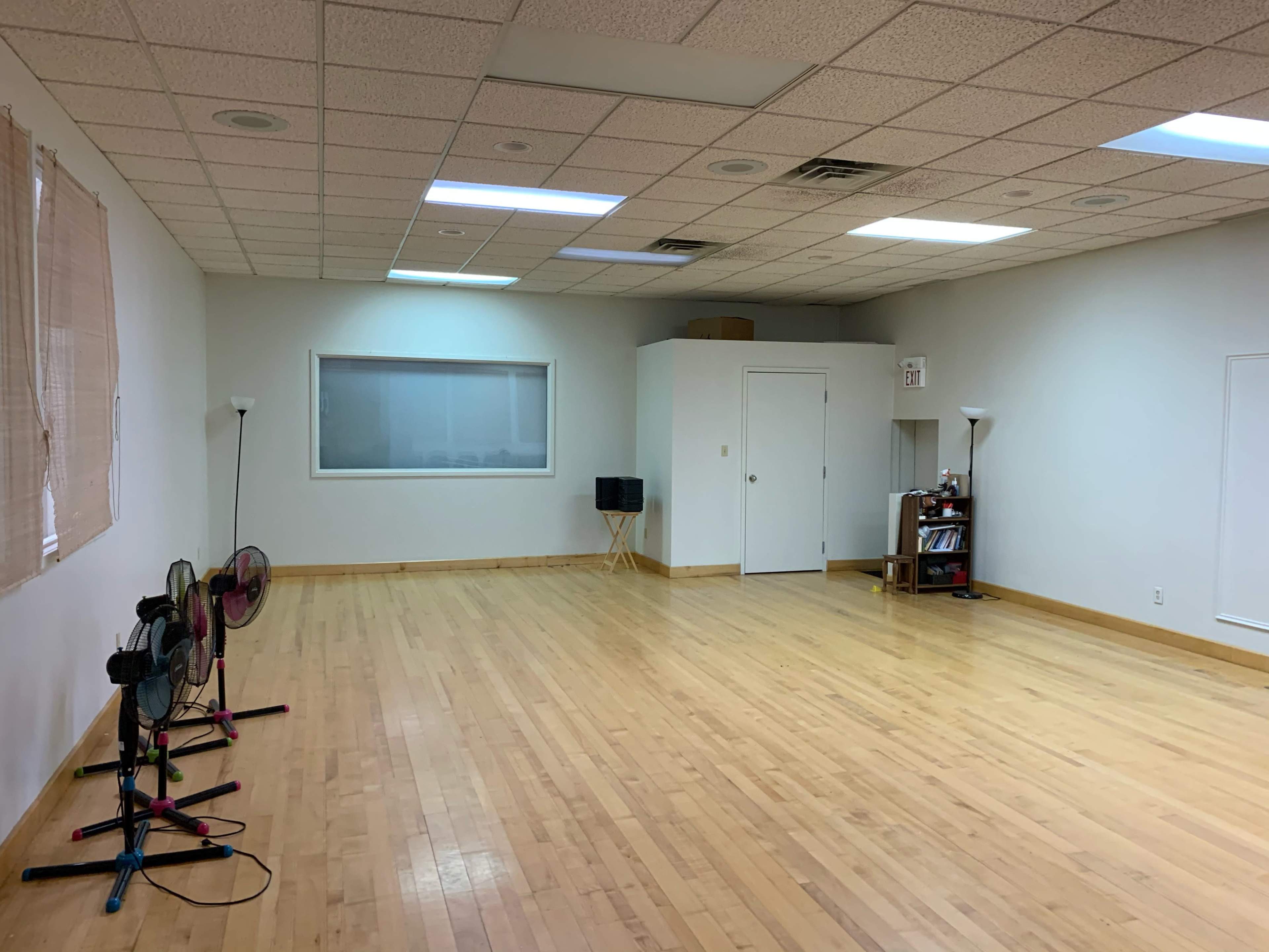 An empty room with wooden floors, a large window, and two standing fans positioned against one wall.