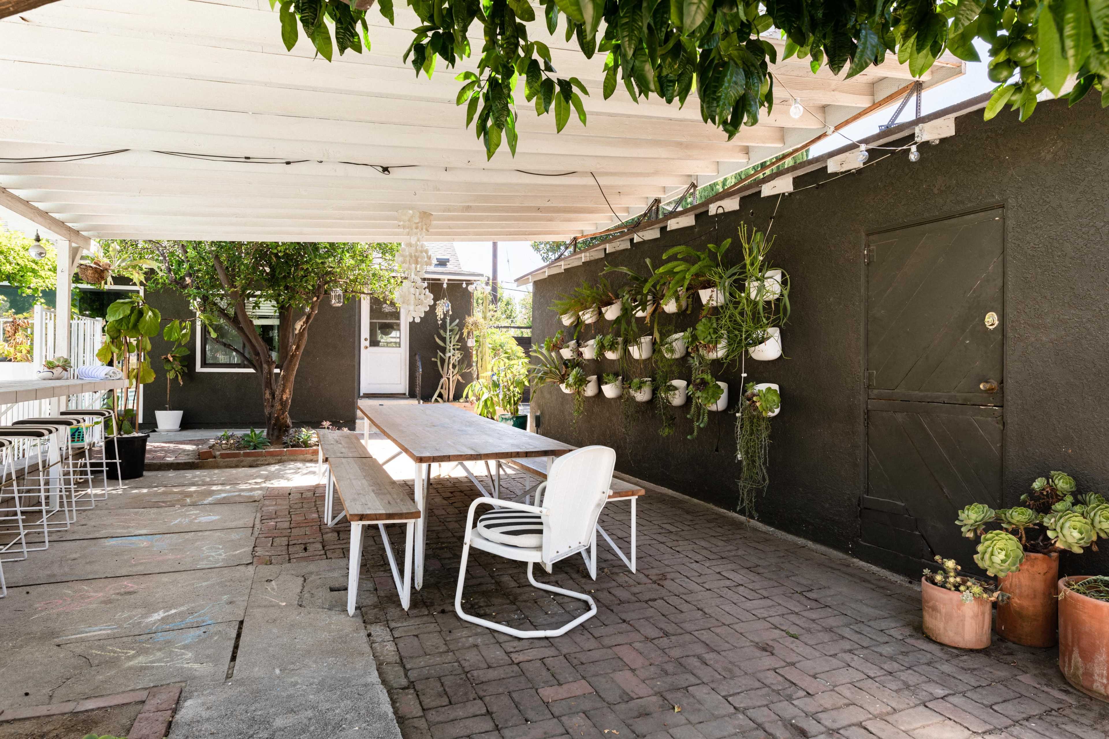 The image shows a shaded outdoor patio area with a wooden table and chairs, surrounded by plants and a wall-mounted vertical garden.