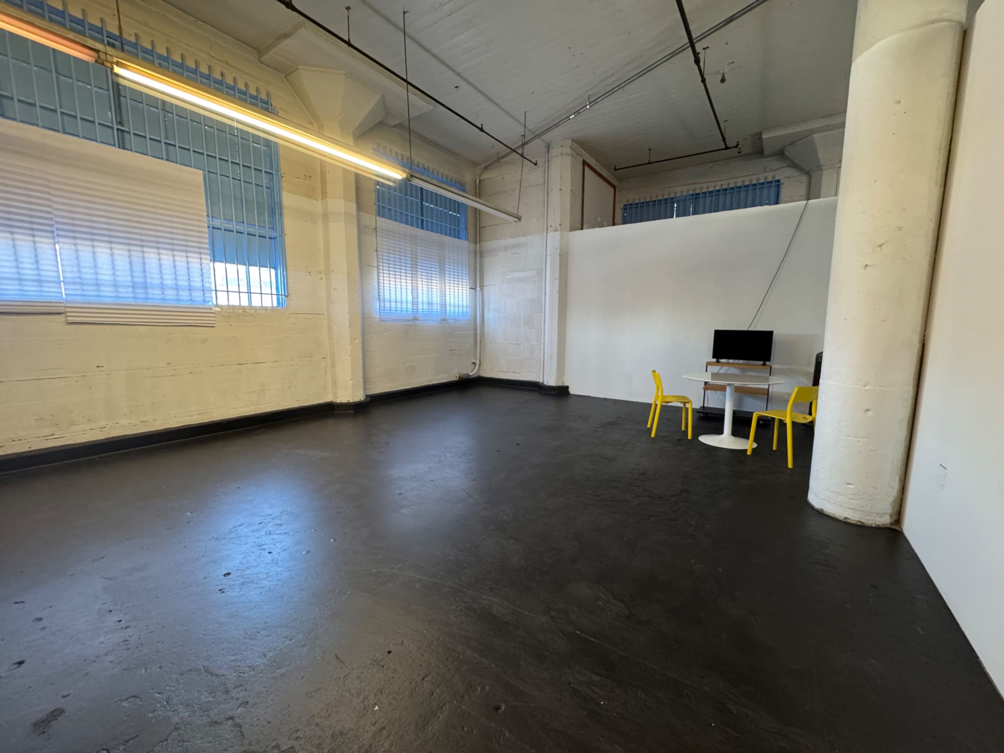 The image shows a spacious, empty room with a black floor, white walls, and a small workspace featuring a table and yellow chairs in one corner.
