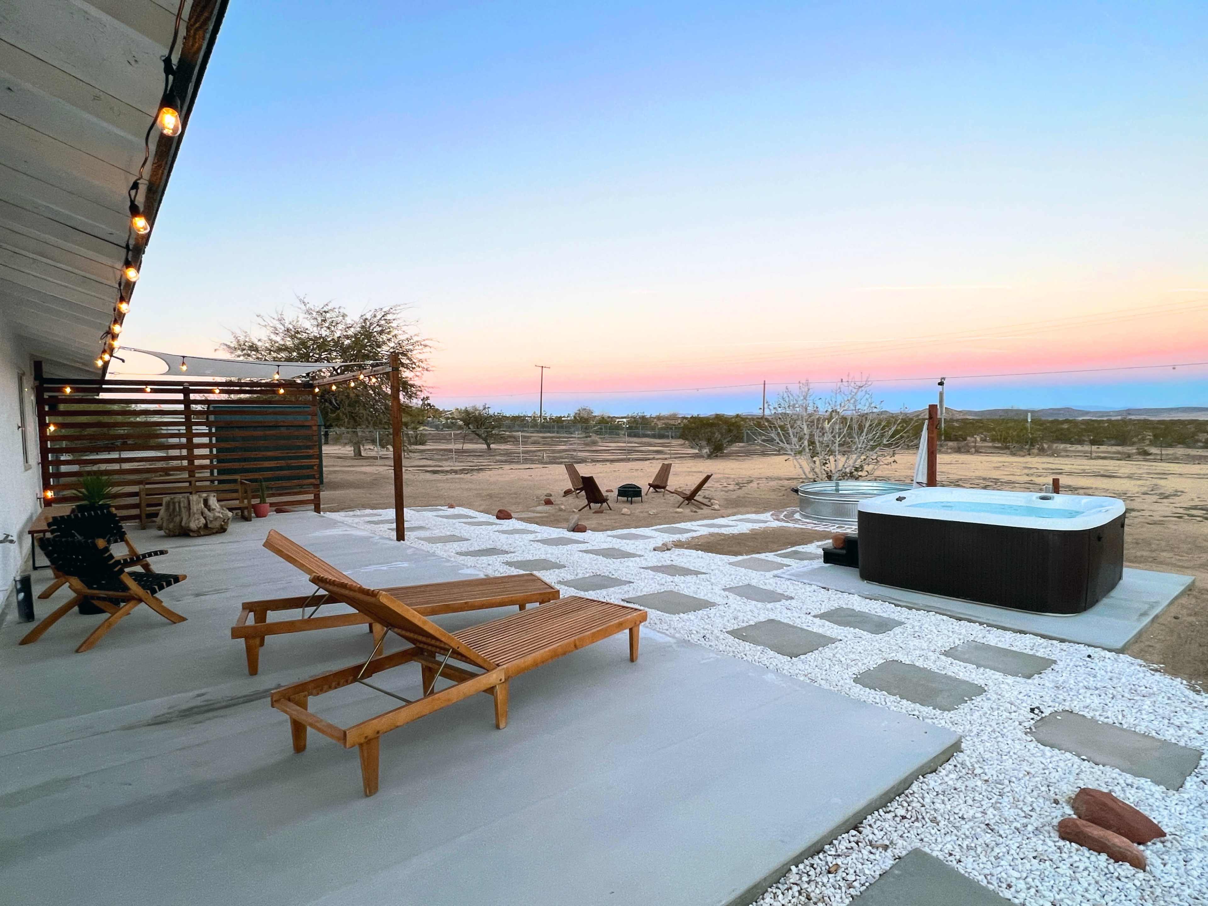 The scene shows a patio area with wooden lounge chairs, a hot tub, and a desert landscape under a pastel sunset sky.