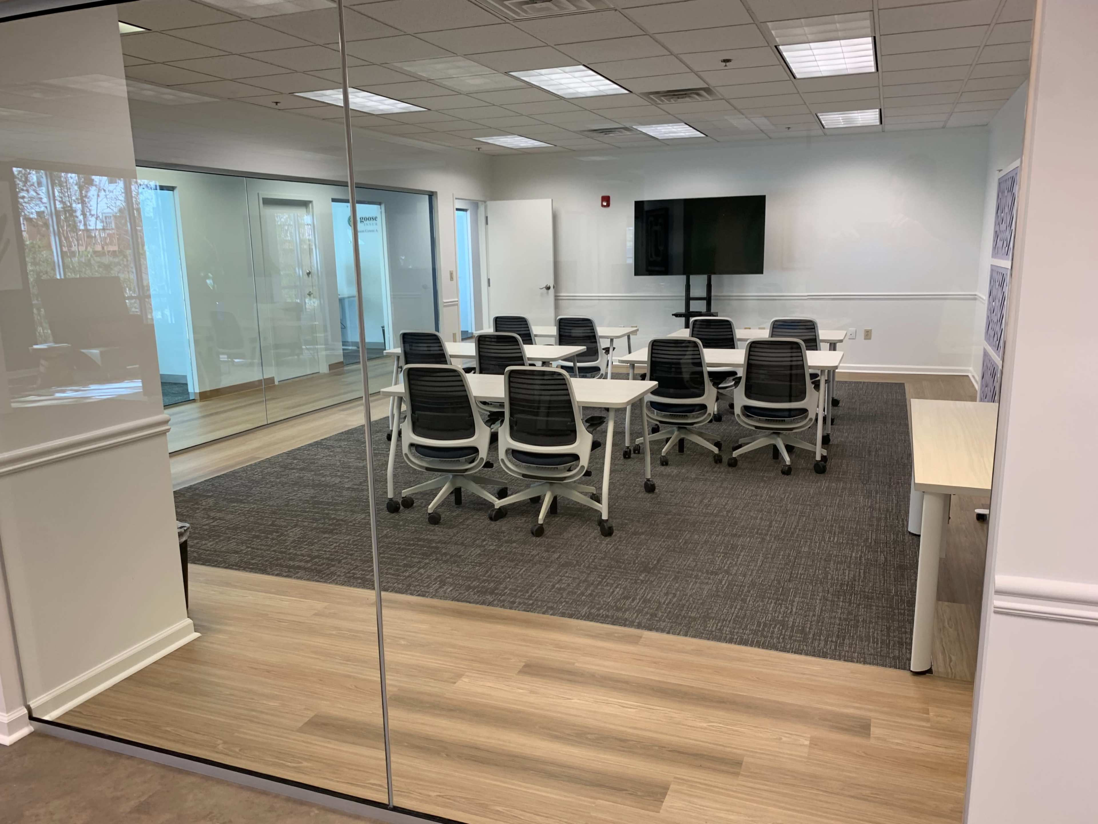 A modern conference room features a large table surrounded by ergonomic chairs, with a wall-mounted screen and glass walls.