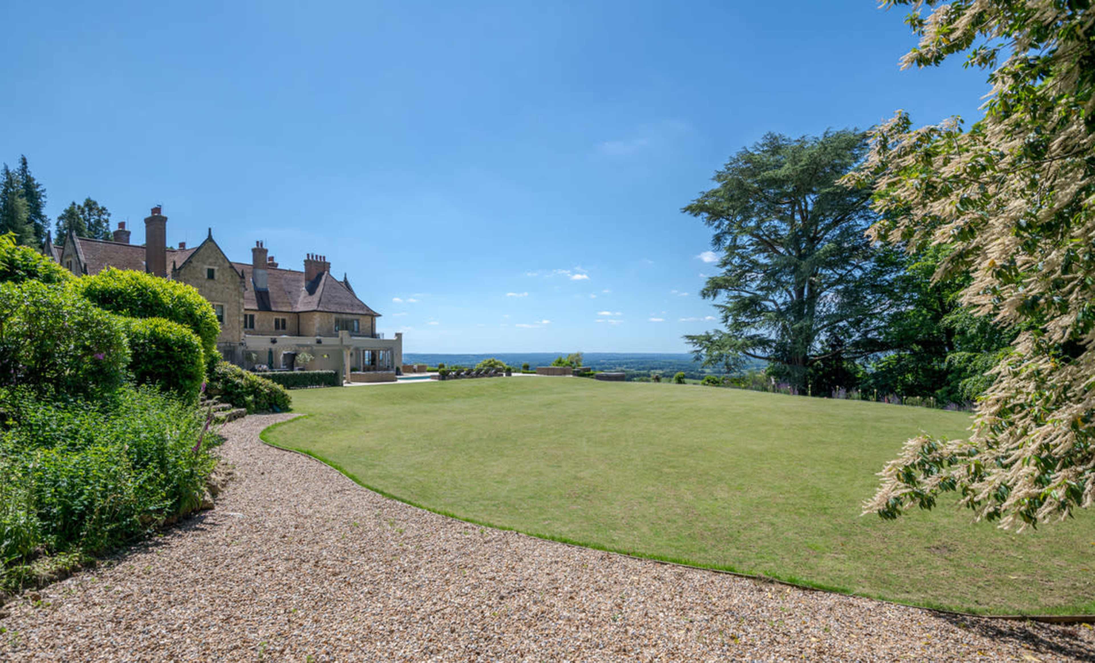 A large mansion with a manicured lawn and a gravel pathway is surrounded by lush greenery under a clear blue sky.