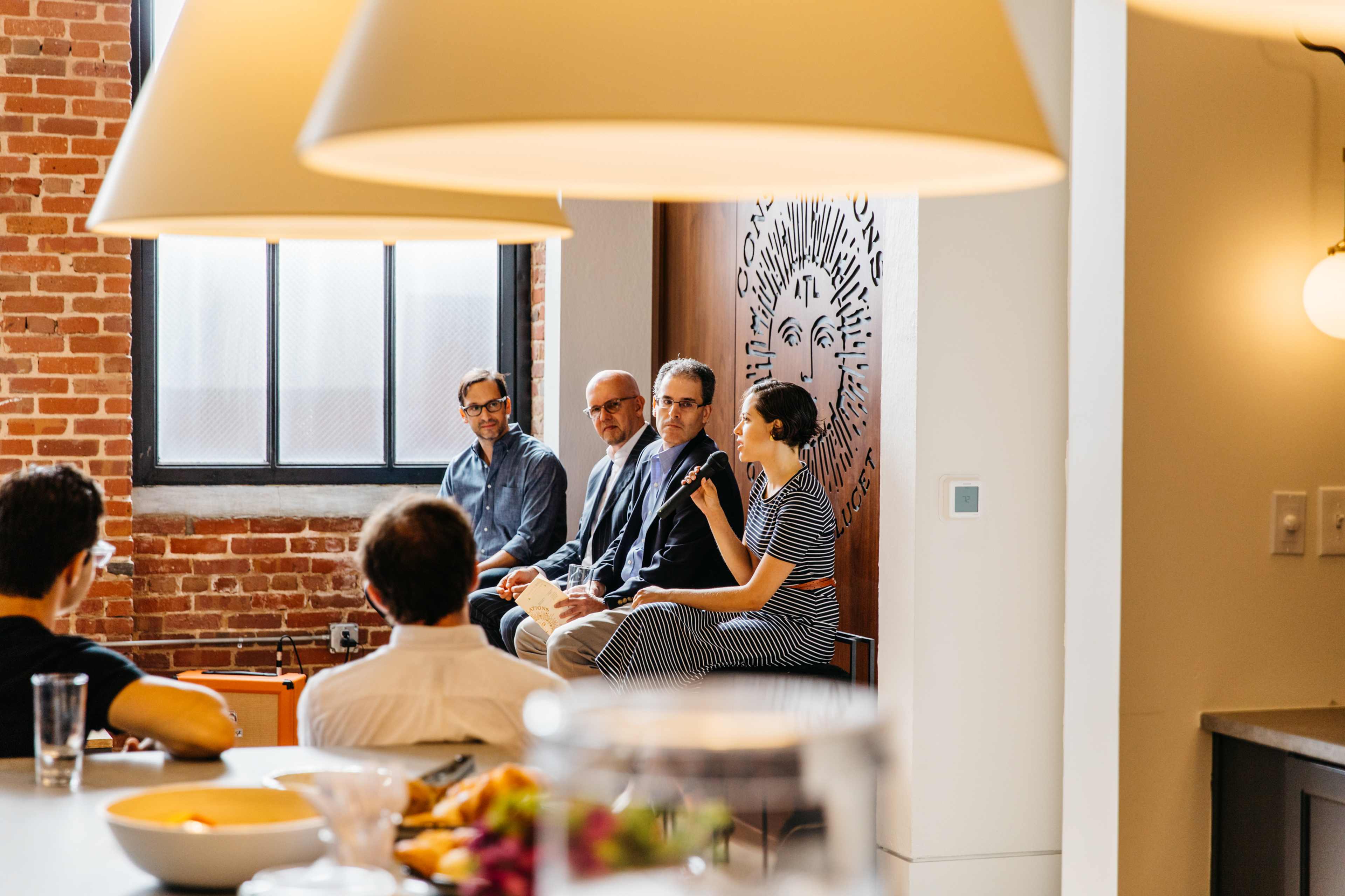 A panel of four speakers is seated in front of an audience in a well-lit room with brick walls and large pendant lights.
